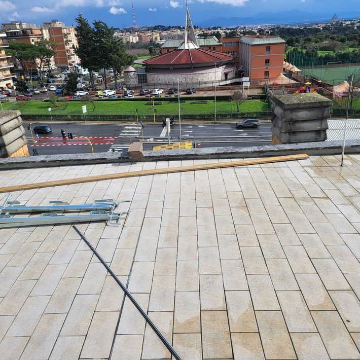Terrazza in pietra affacciata su una strada, un edificio dal tetto rosso e campi verdi sotto un cielo nuvoloso.