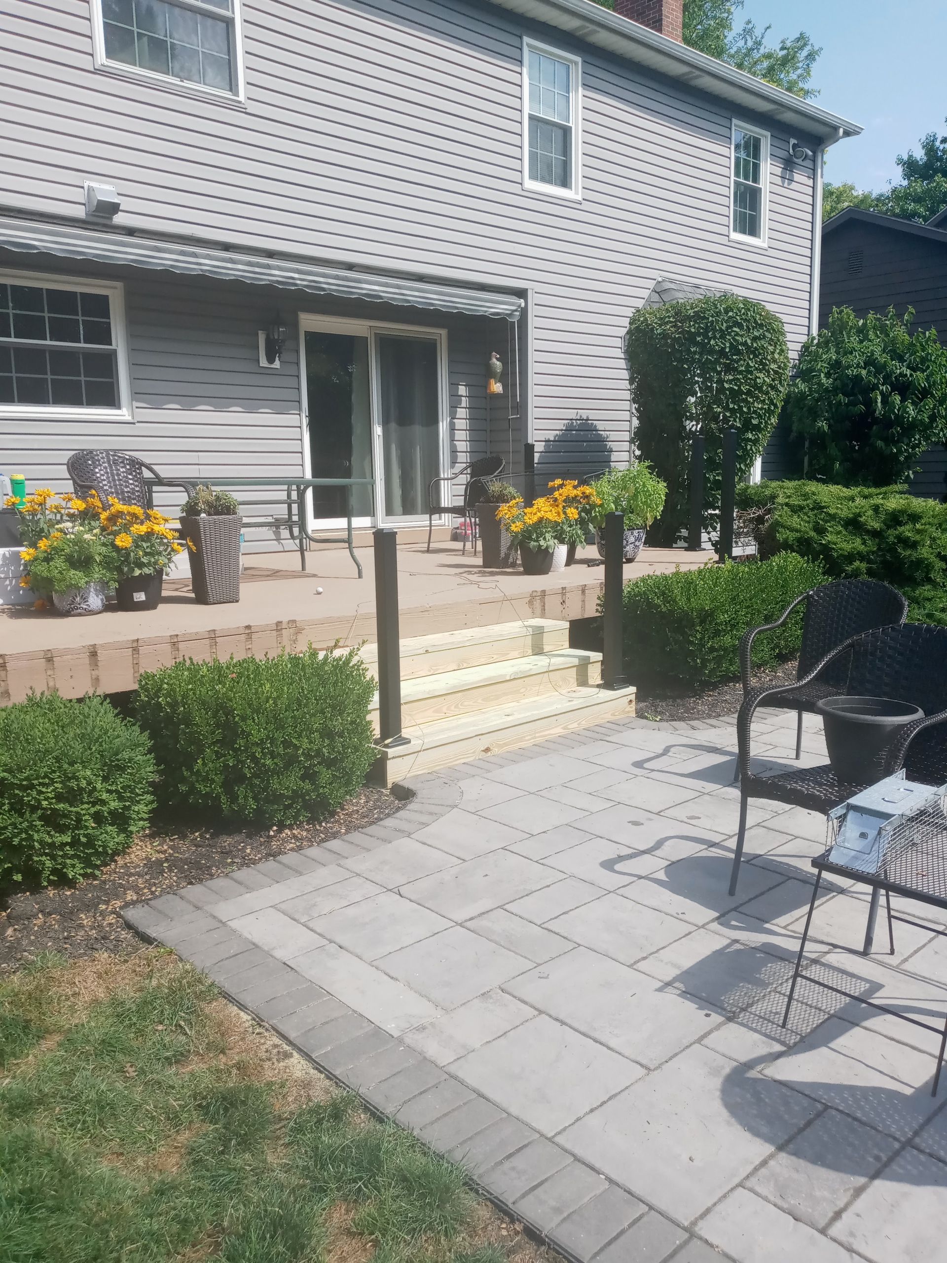 Backyard patio with paved walkway and steps leading to a deck with sliding doors, surrounded by bushes and potted plants.