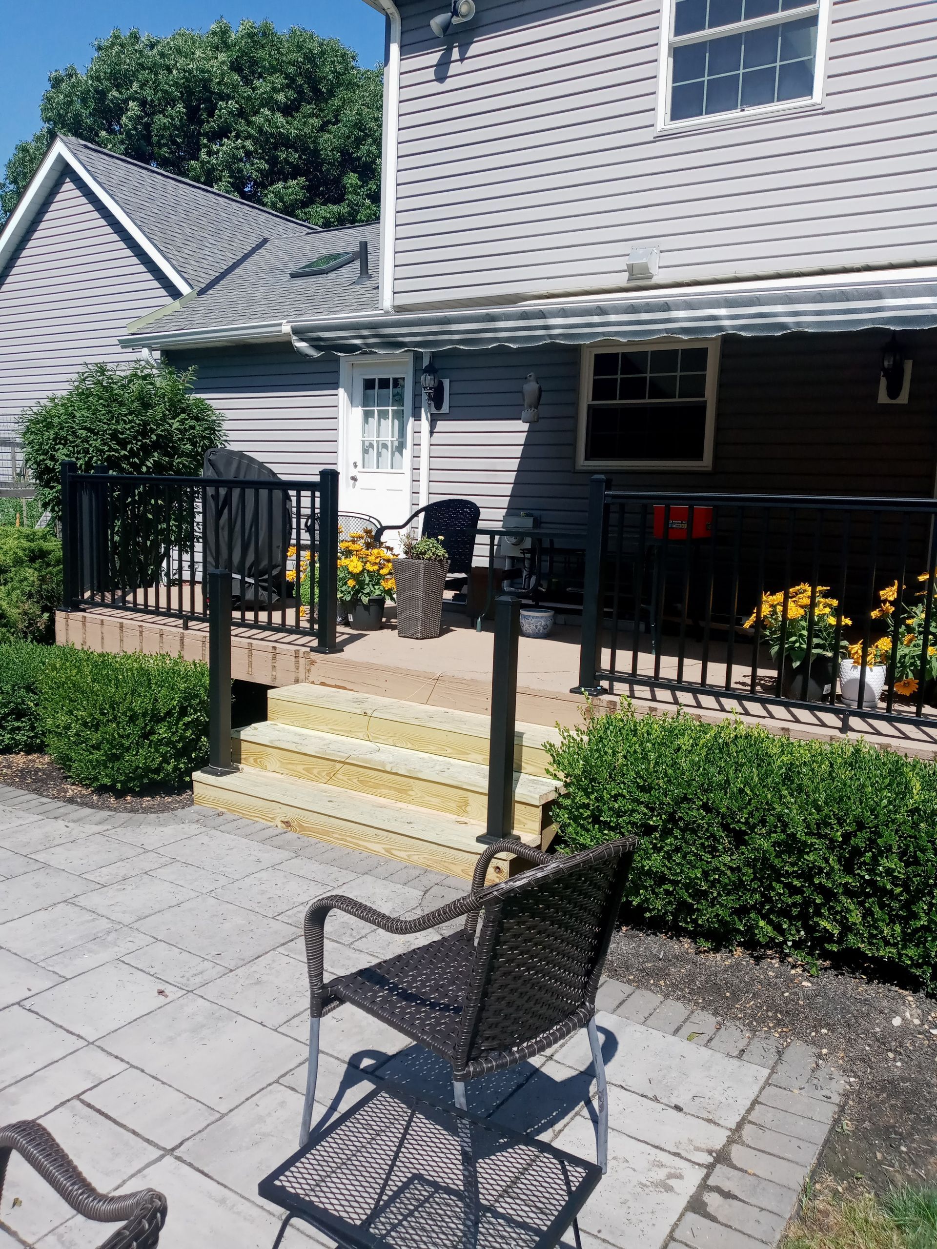 Backyard patio with deck, steps, black railing, and potted yellow flowers. Wicker chair in foreground. Gray house.
