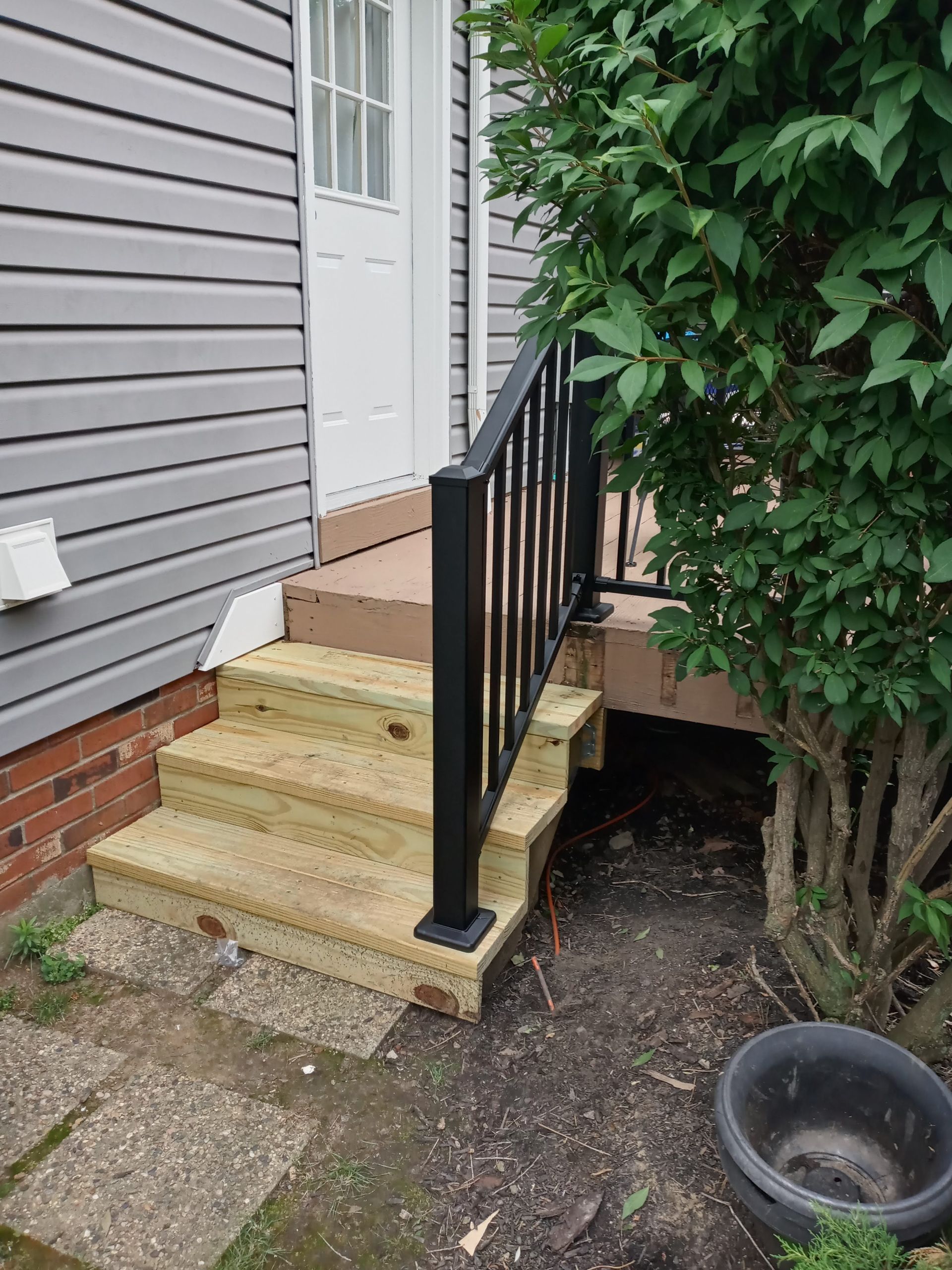Wooden stairs leading to a doorway, with a black handrail and a bush nearby.