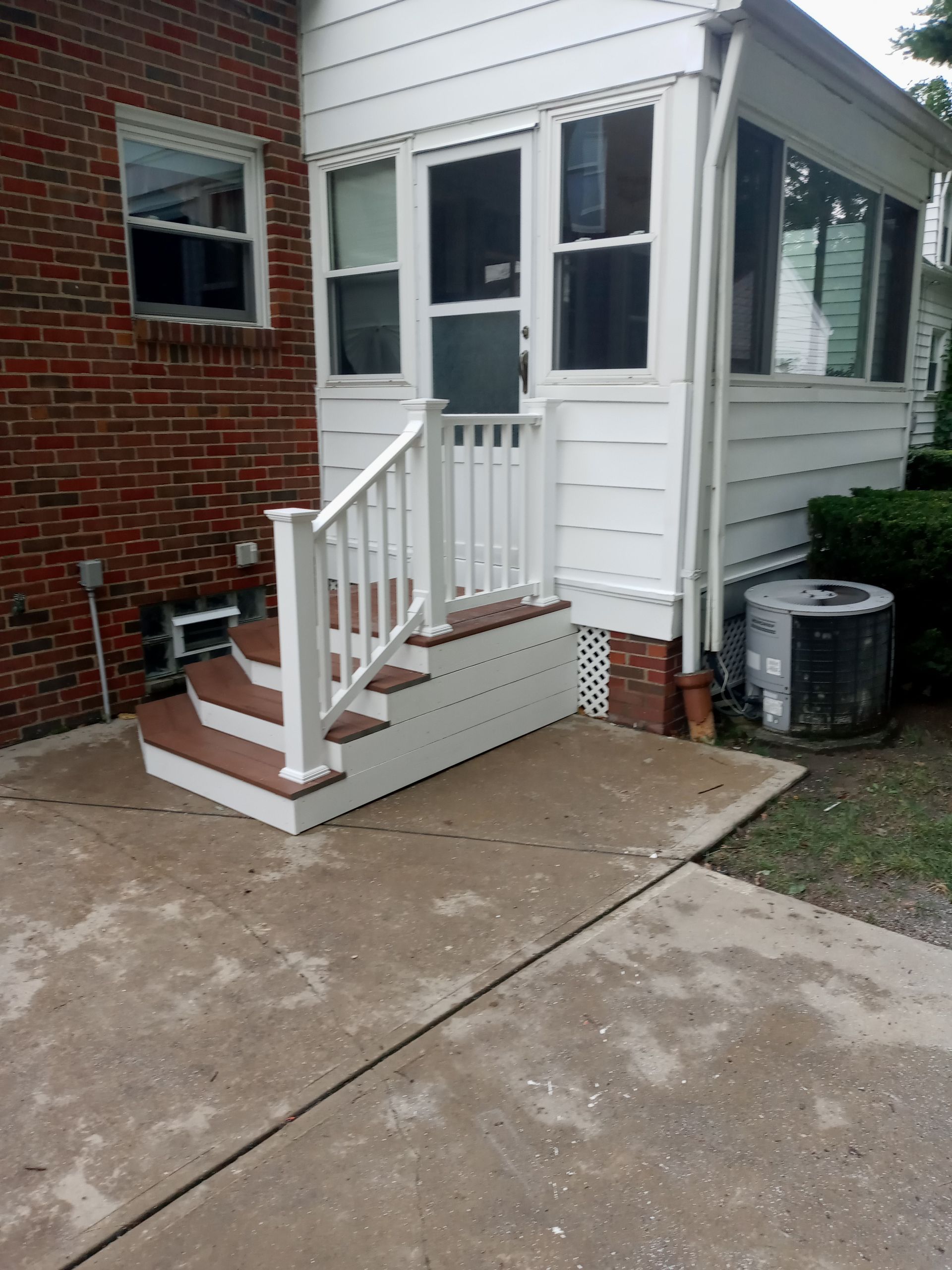 Brick house with a composite deck staircase and white railing leading to a white-sided porch. Concrete patio.