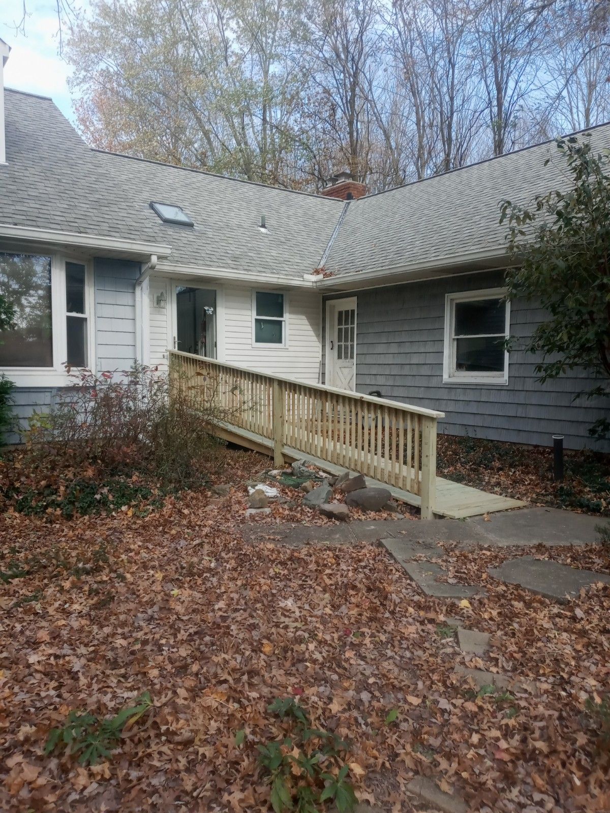 Ranch-style house with wooden wheelchair ramp. Fall leaves cover ground. Gray siding, white doors, and windows.