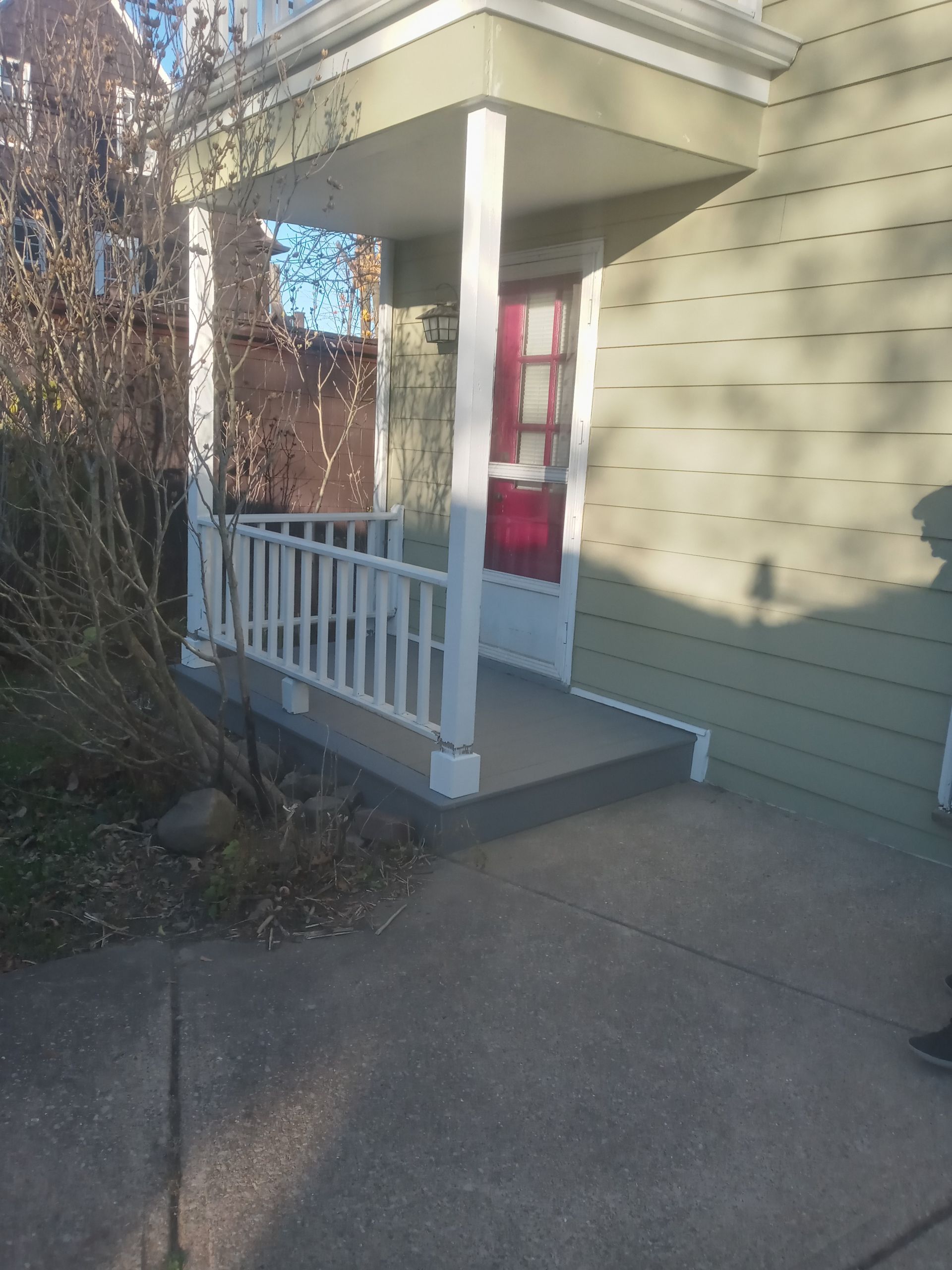 Small front porch with white railing and red door, next to a pale green house.