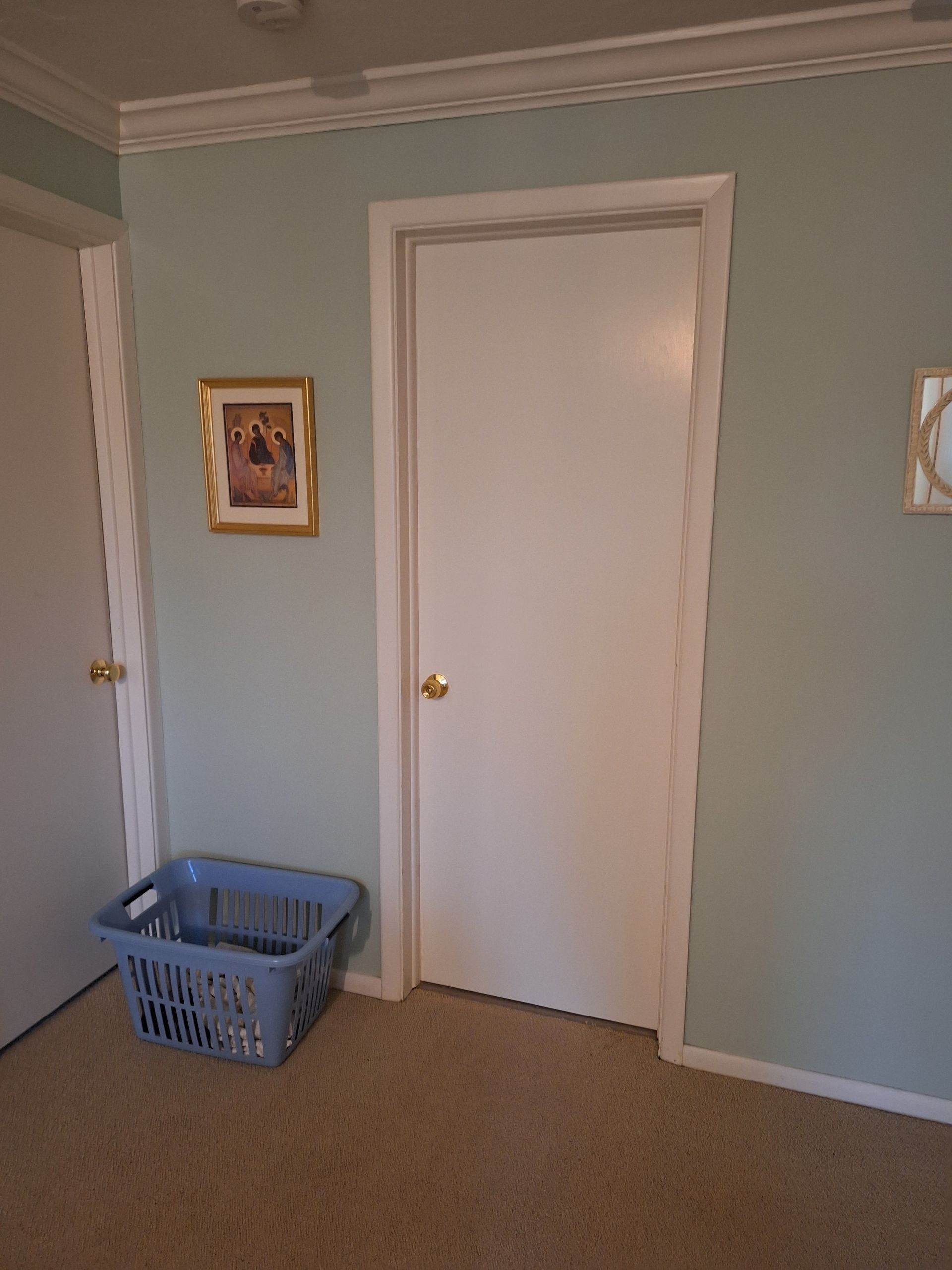 Blue laundry basket near a closed white door in a room with light blue walls and beige carpet.