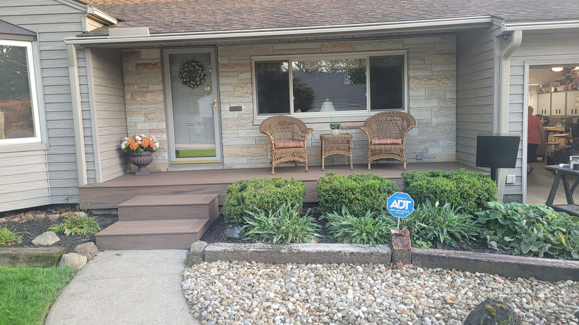 A small beige house with a porch and two wicker chairs, shrubs, and a security system sign.