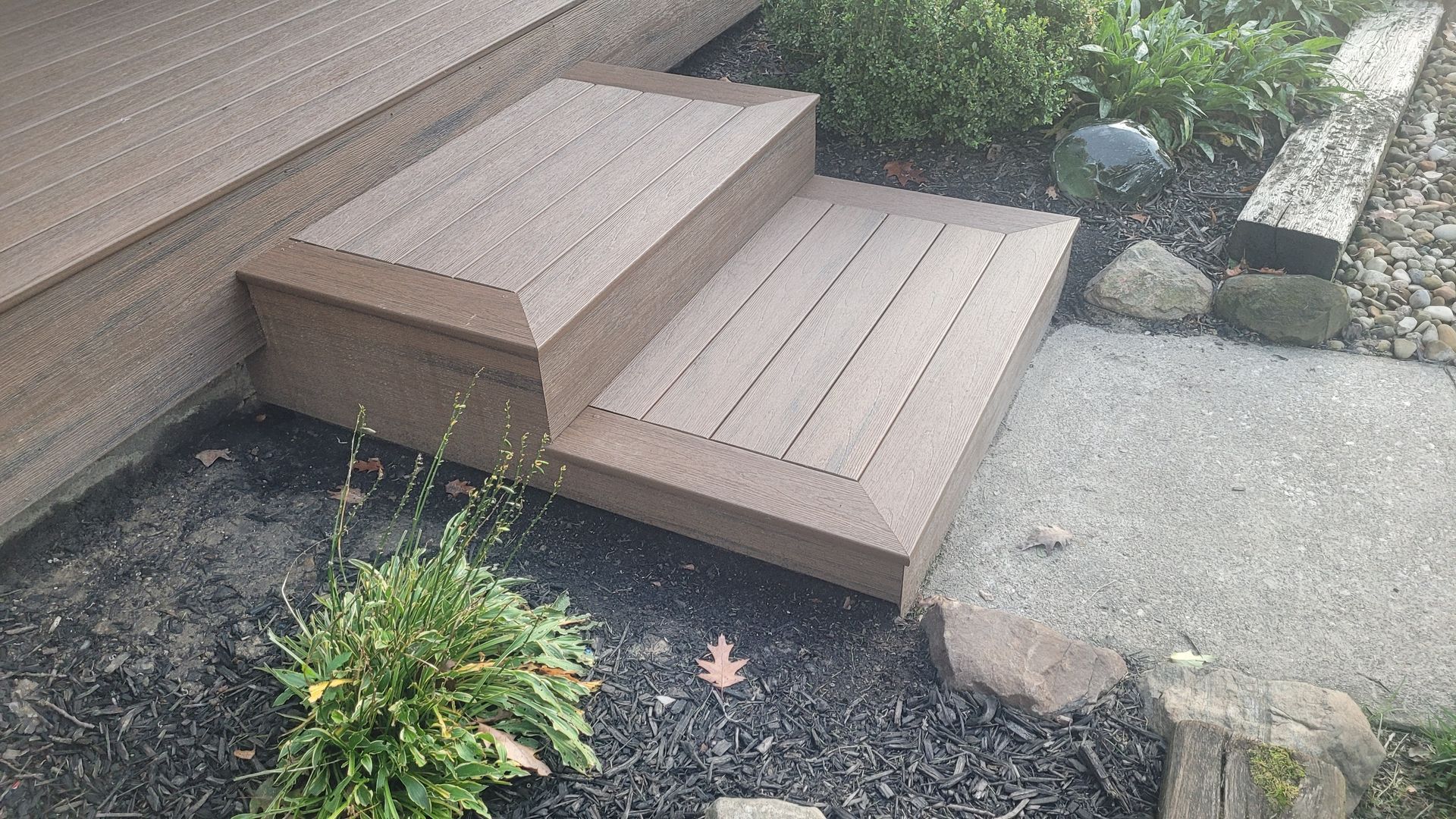 Brown composite deck steps leading from a raised platform to a gravel walkway, surrounded by landscaping.