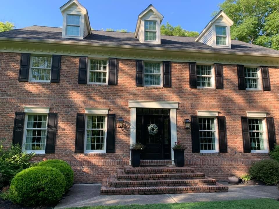 Red brick house with black shutters, white trim, and a black door; shrubs in the foreground.