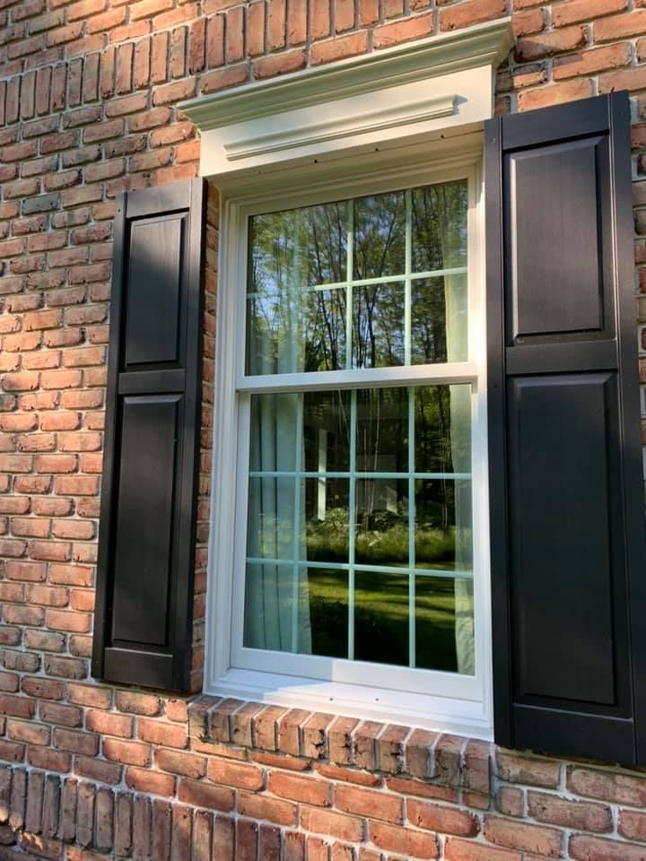 Black shutters flank a white-framed window with a grid pattern. The window is set in a red brick wall.