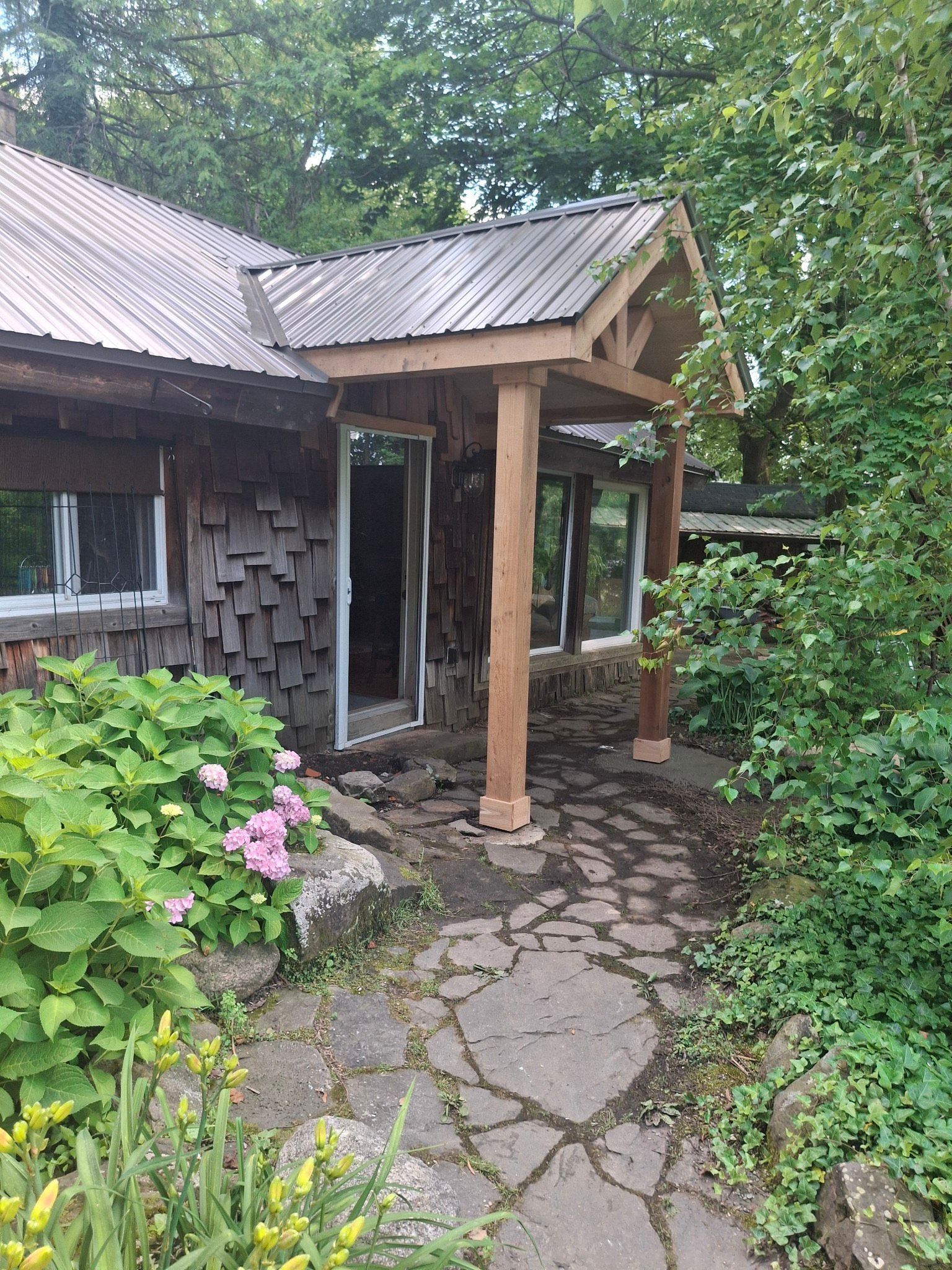 Wooden cabin with a stone path leading to a covered entrance and glass doors, surrounded by greenery.