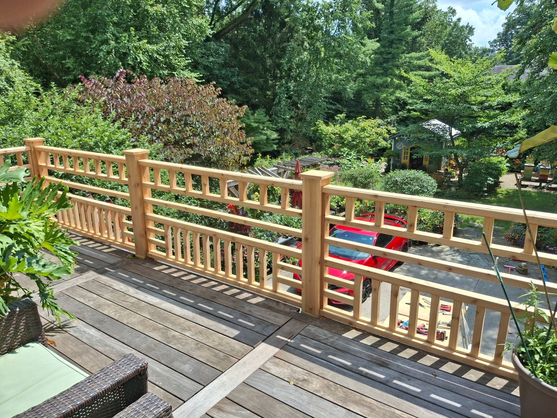 Wooden deck with decorative railing, trees and shrubs in the background. Sunny day.
