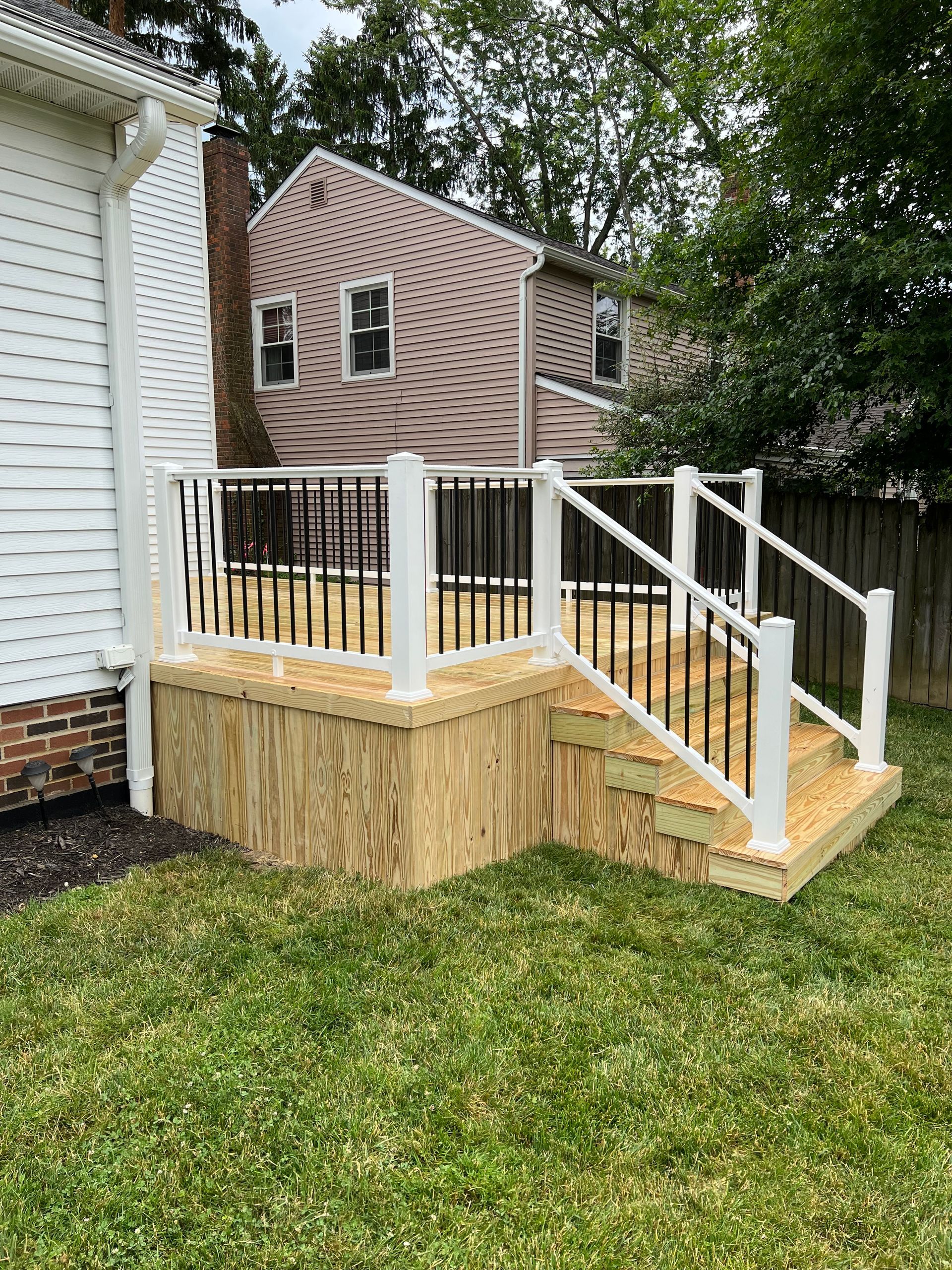 Wooden deck with white railing, black spindles, and steps, next to a white house and green grass.