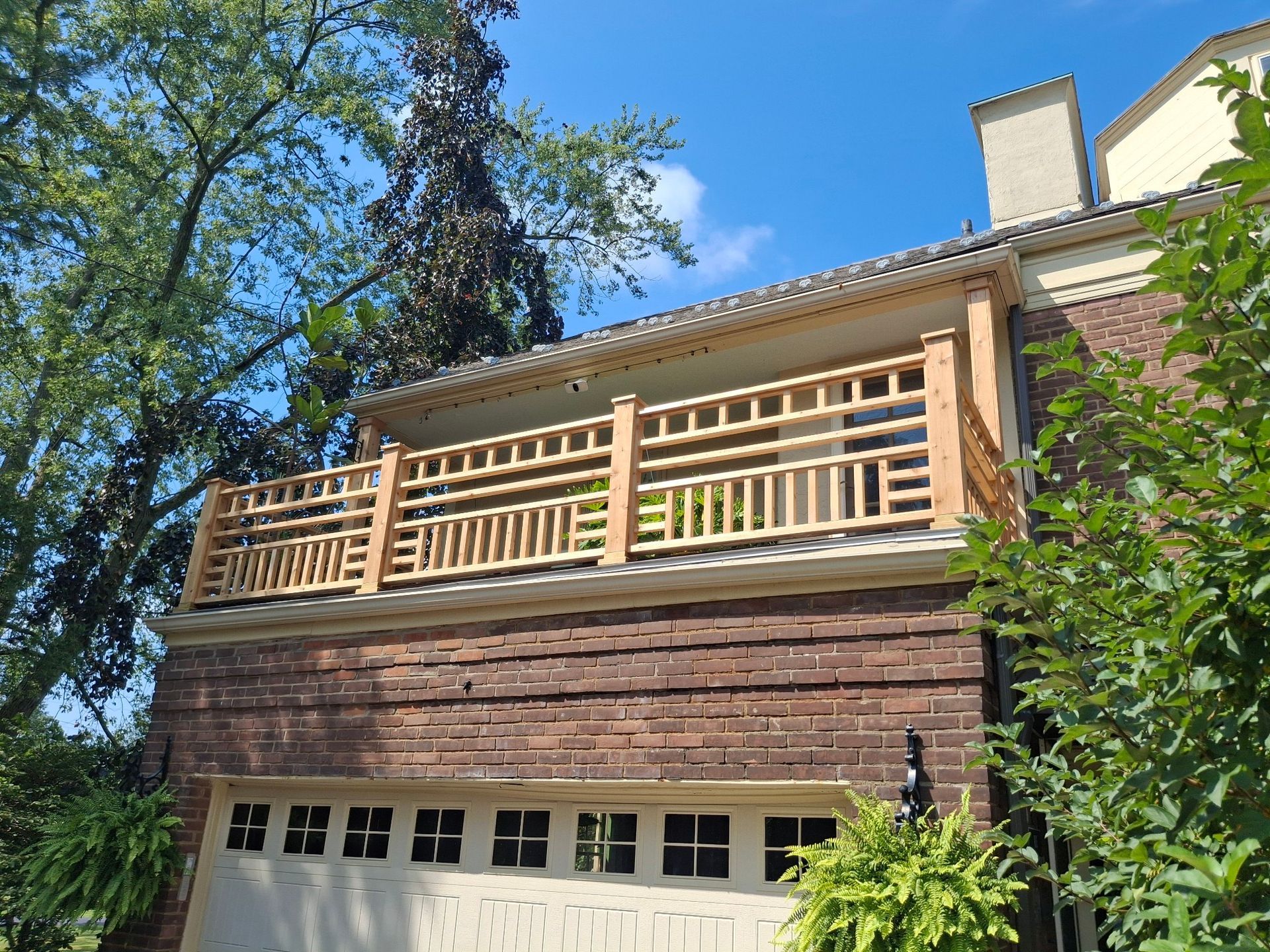 Wooden balcony with decorative railing above a brick garage, sunny day.