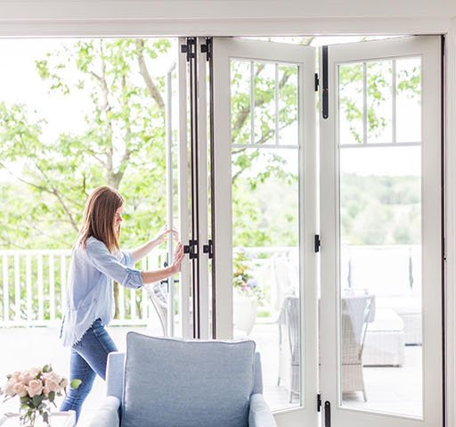 Woman opening folding glass doors onto a patio with furniture and green trees visible.