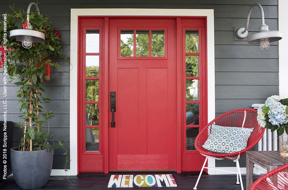 Red front door with sidelights and welcome mat on a gray porch, with a chair and potted plant.