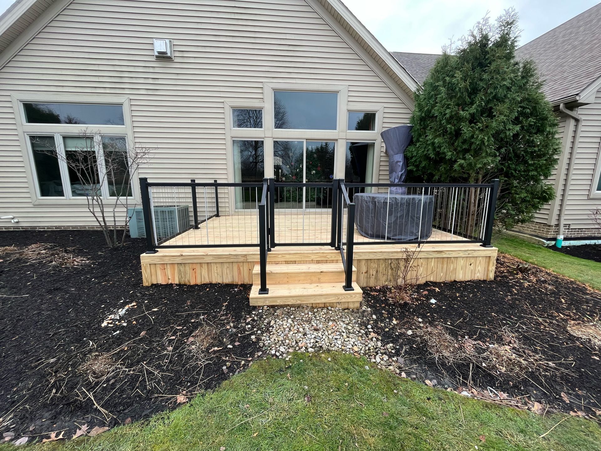 Wooden deck with black railing and steps in front of a house, surrounded by mulch and grass.
