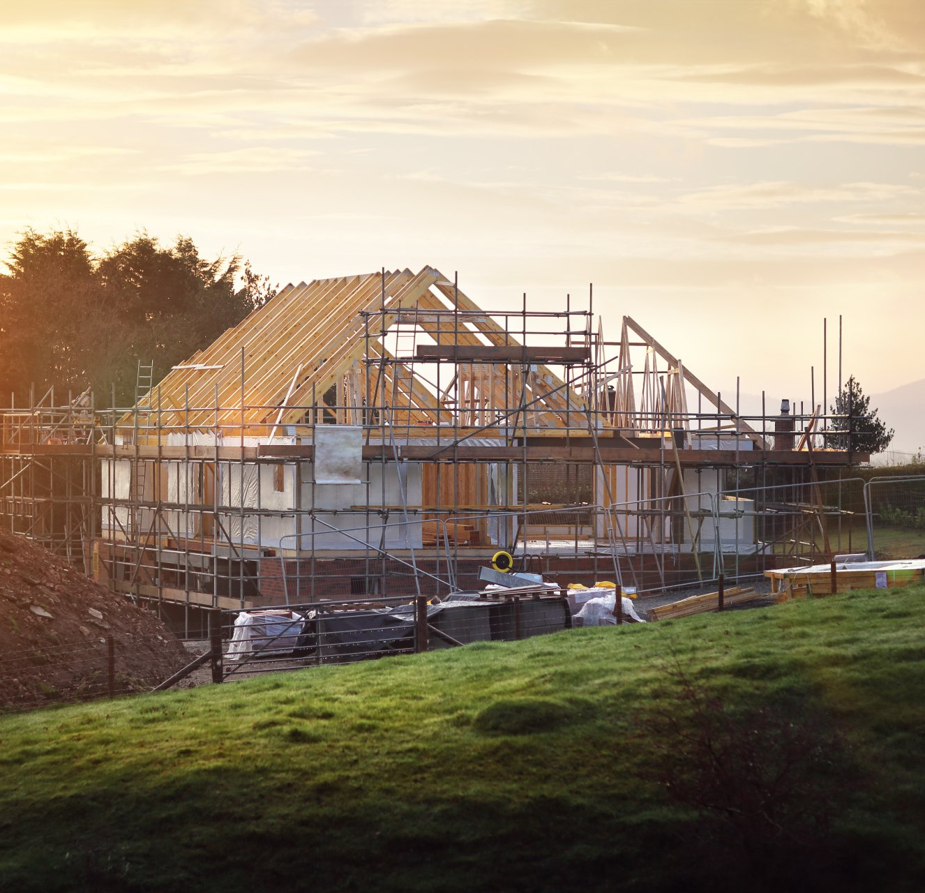 House under construction with exposed wooden frame and scaffolding on a grassy hill at sunset.