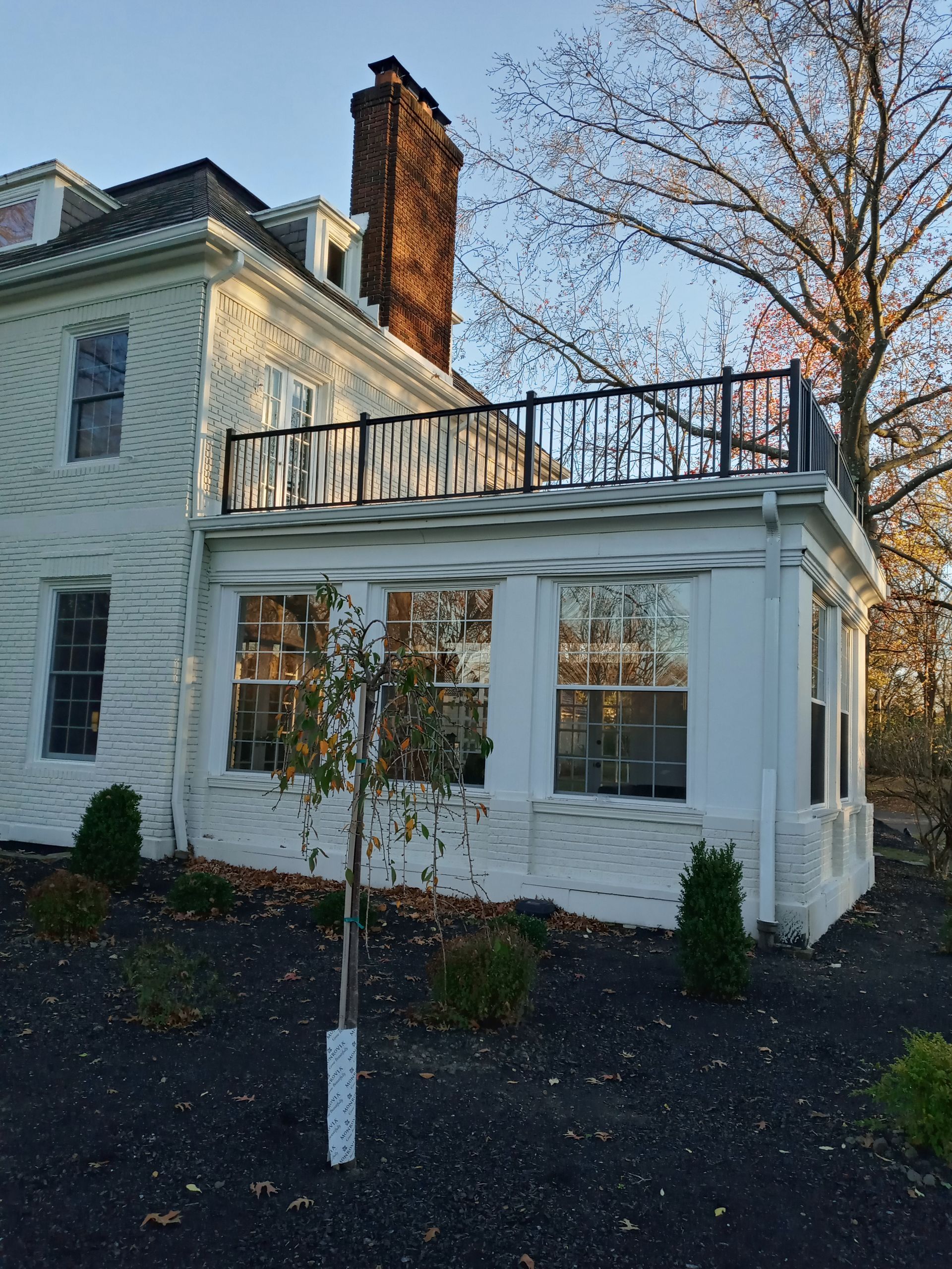 White house with a sunroom, small tree in front, a rooftop deck with black railing.