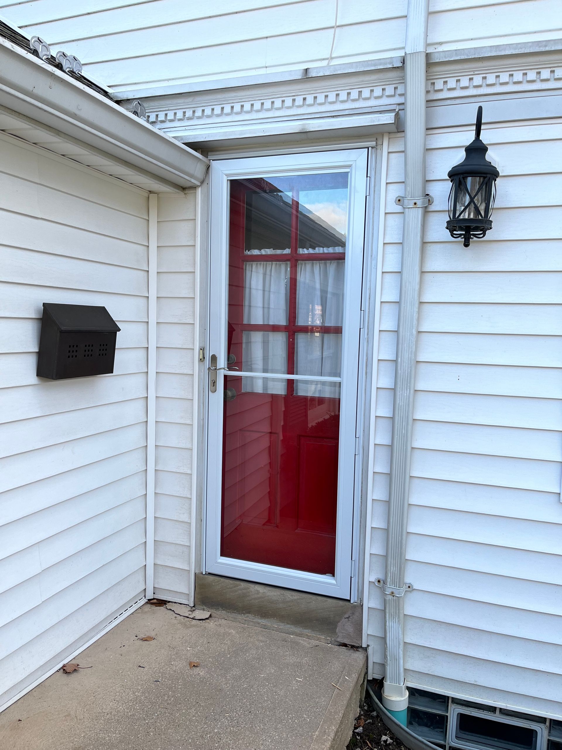 Red door with storm door, white siding, black mailbox, and wall-mounted lamp.