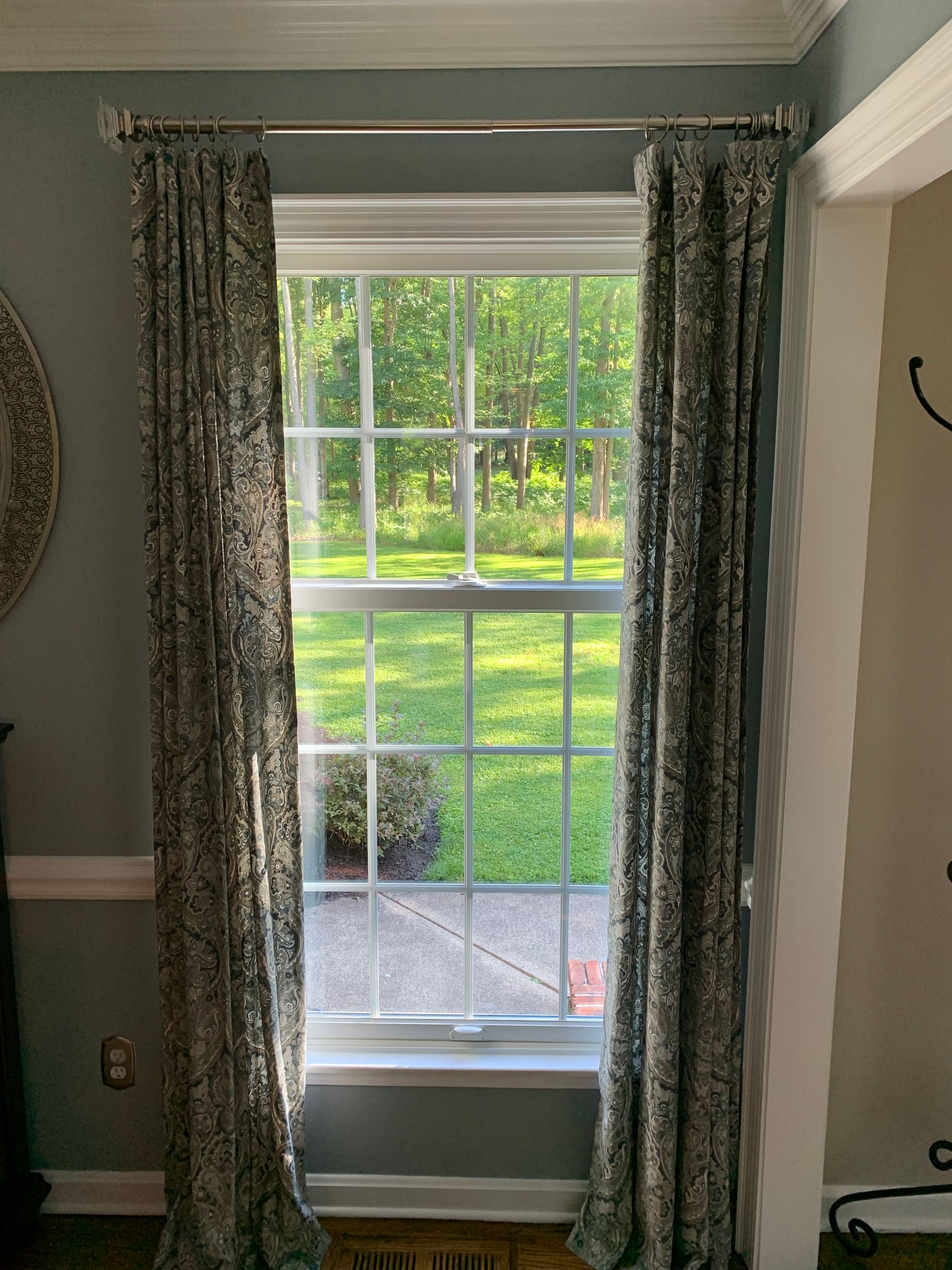 Window with patterned drapes, looking out onto a grassy yard and trees. White trim and frame.