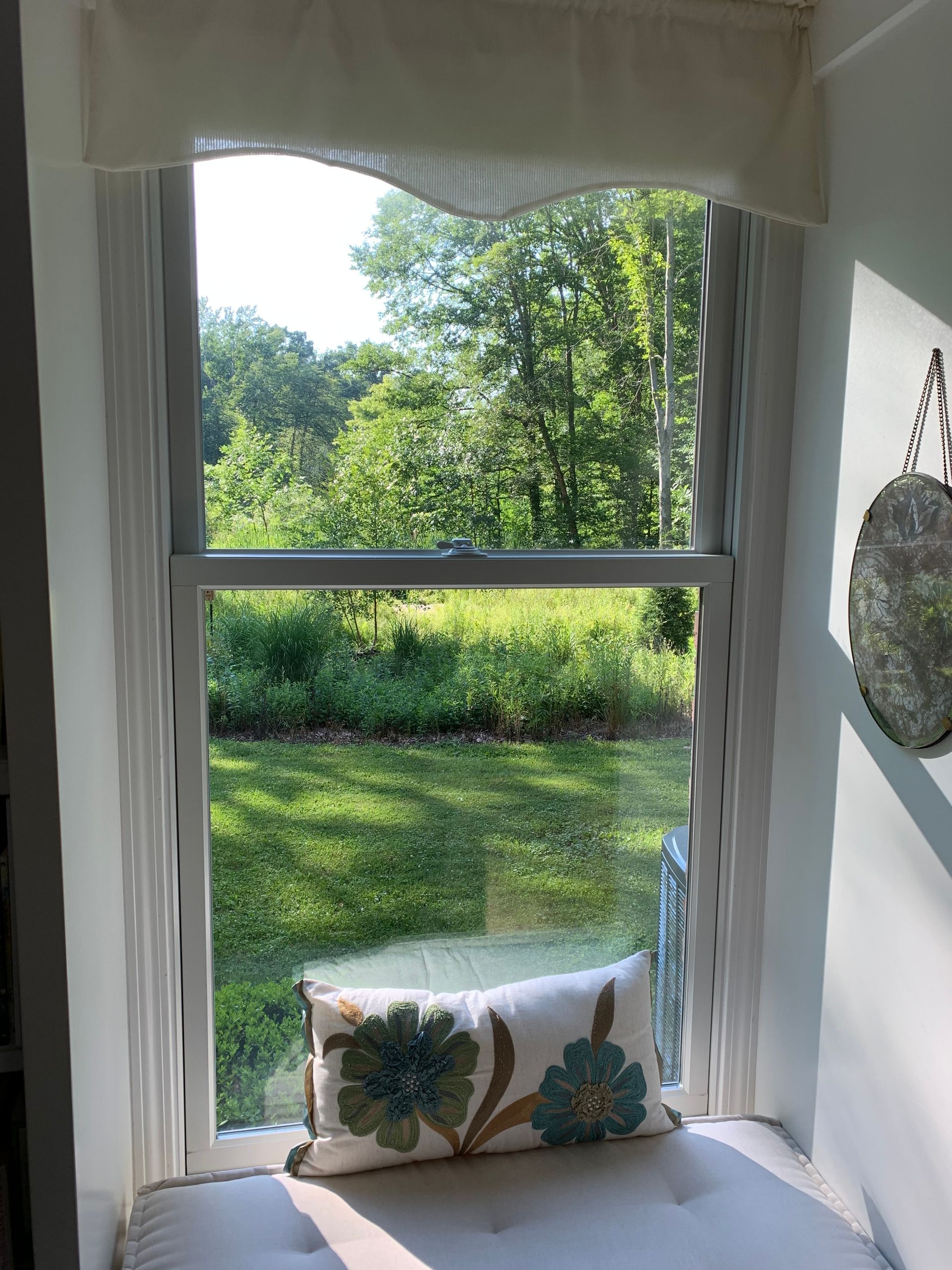 Window seat with view of greenery, pillow, and cream-colored shade.