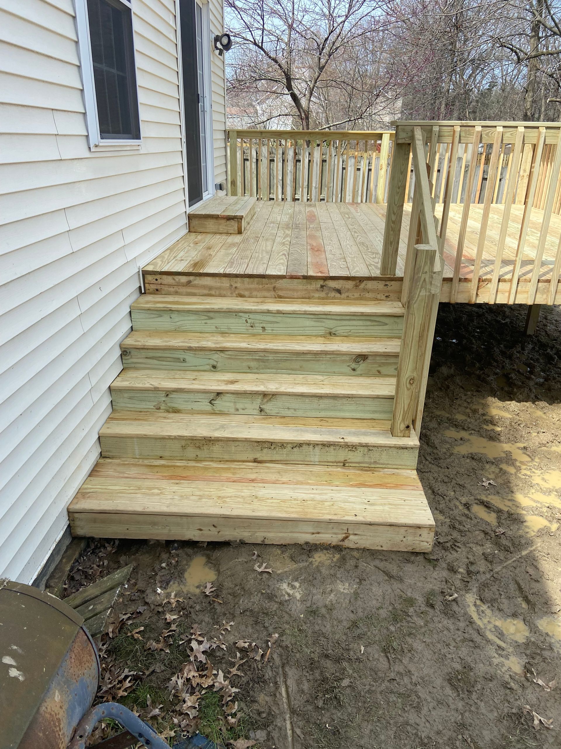 Wooden deck with steps leading to a house entrance. Newly built with light wood, set in a muddy yard.