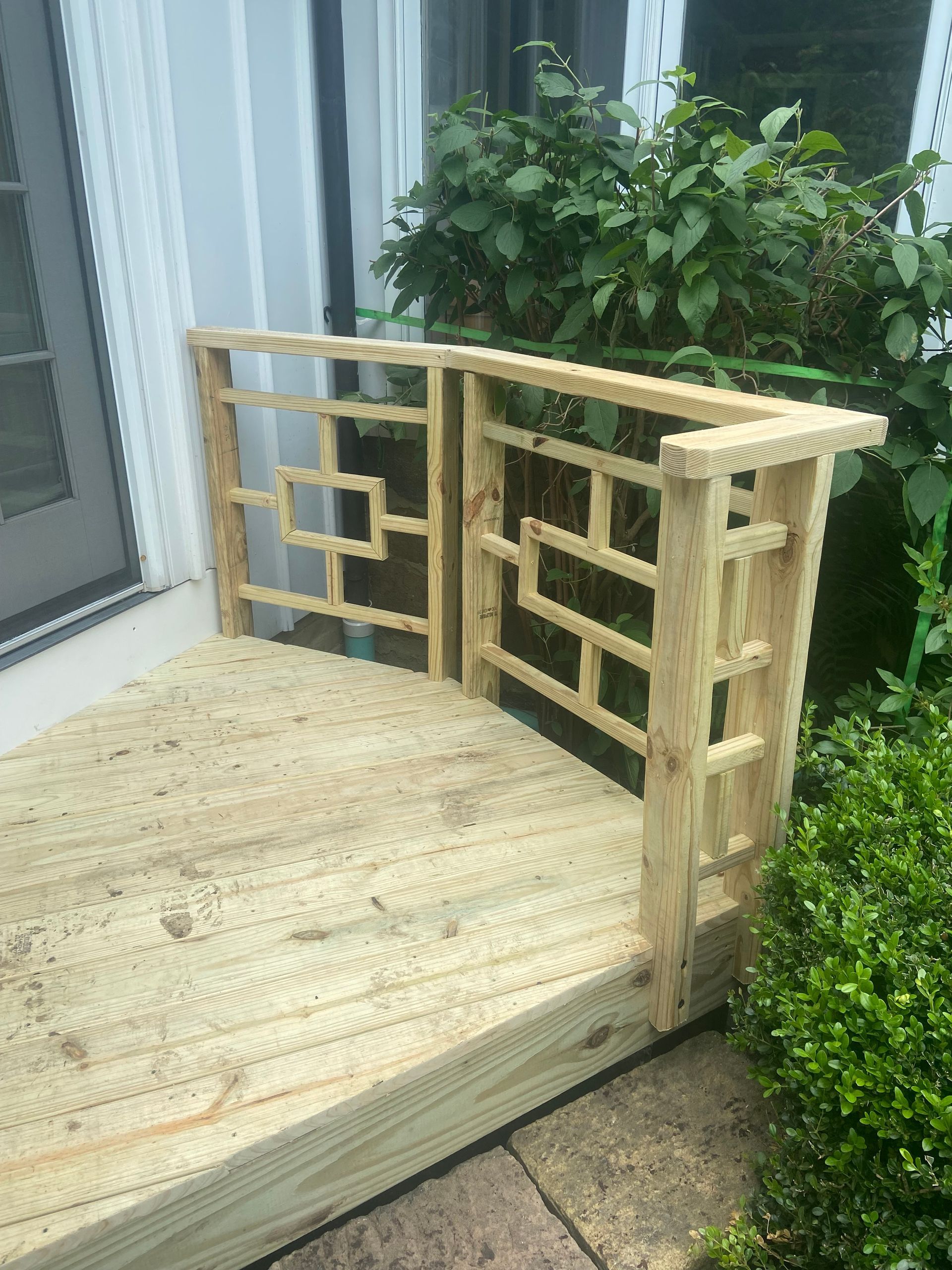 Wooden deck with decorative railing beside a door, next to greenery.