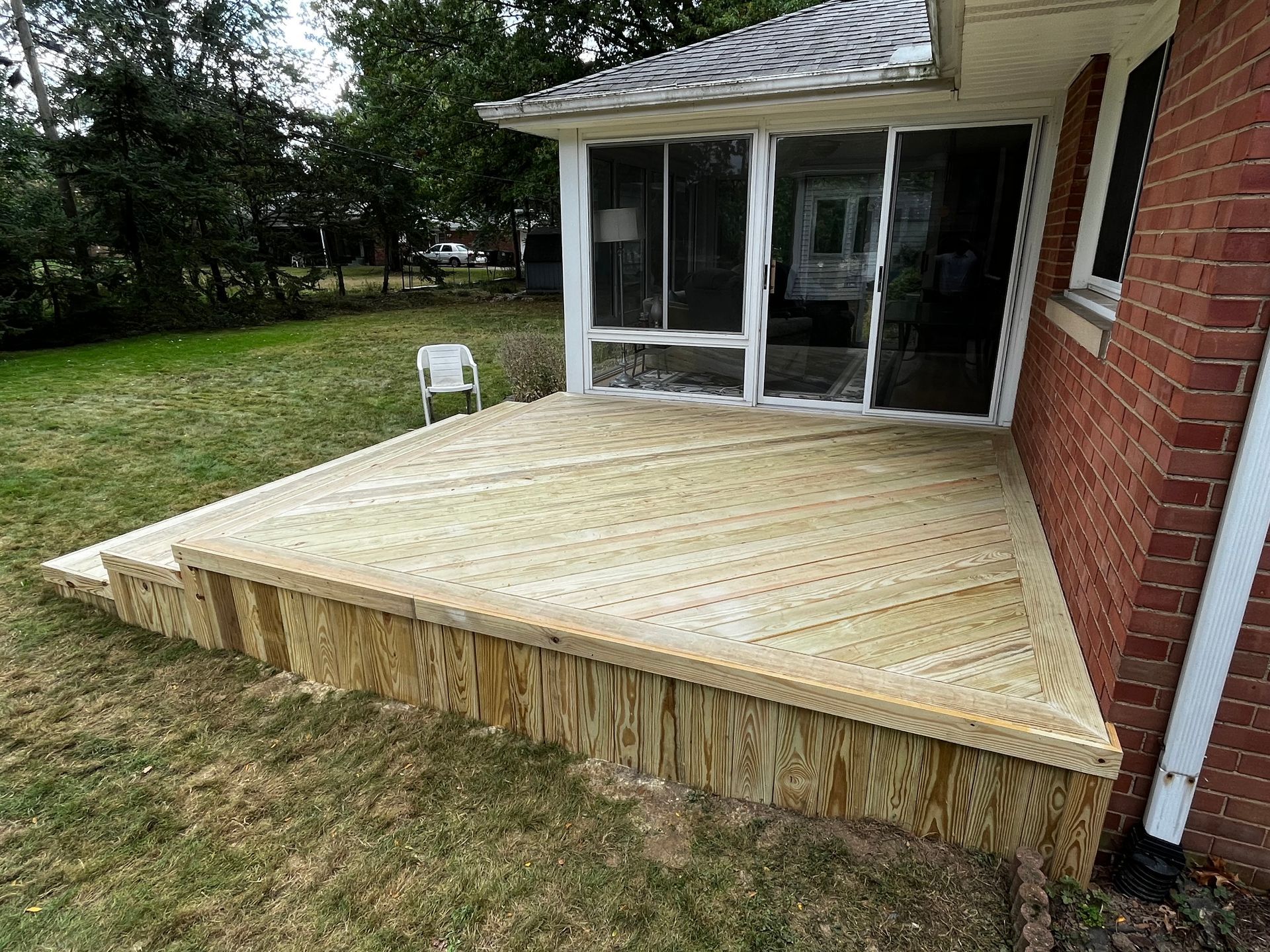 Wooden deck with steps next to a brick house and screened-in porch, surrounded by grass.