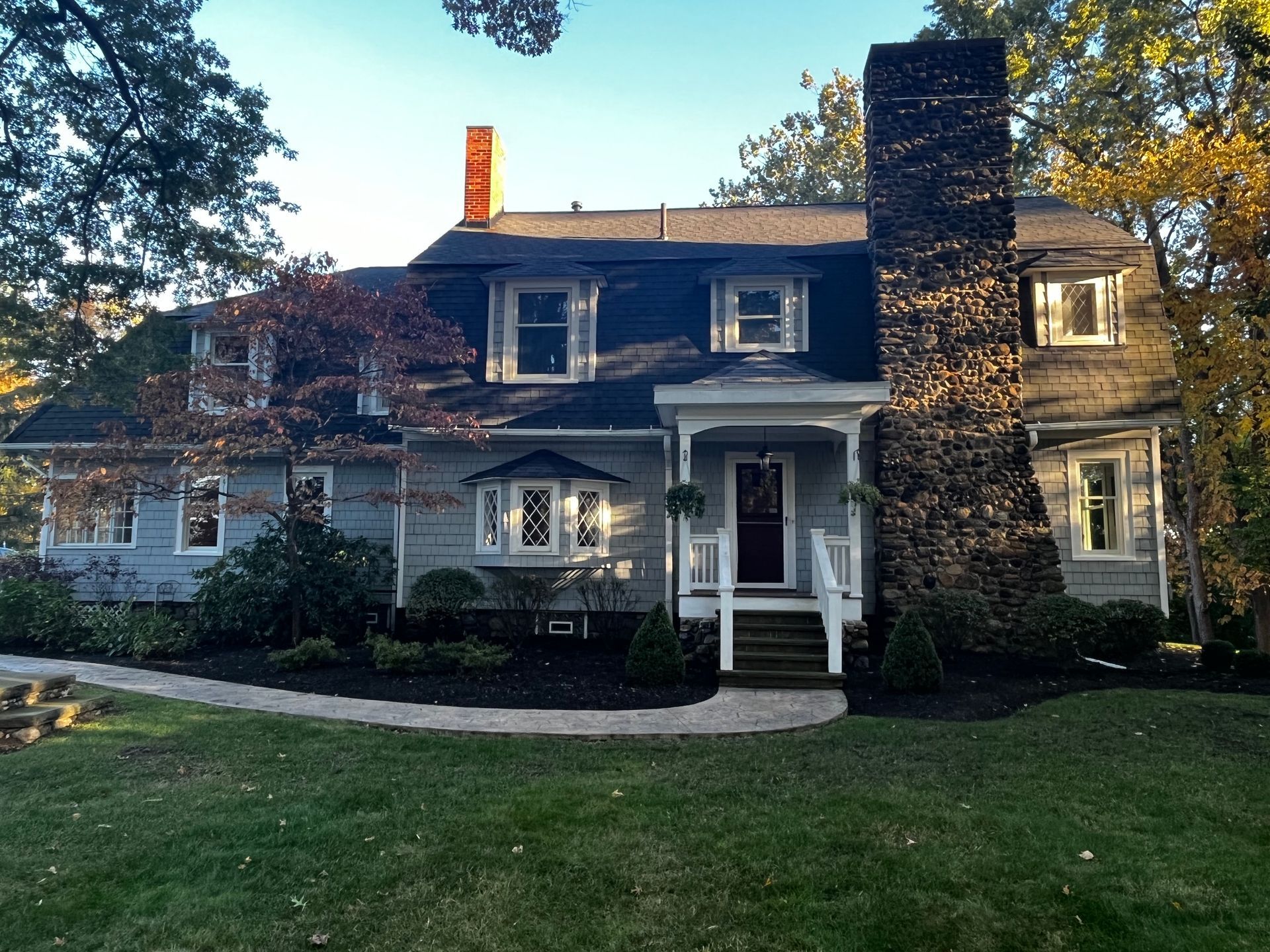 Two-story gray house with a stone chimney, dark roof, and small front porch on a grassy lot.