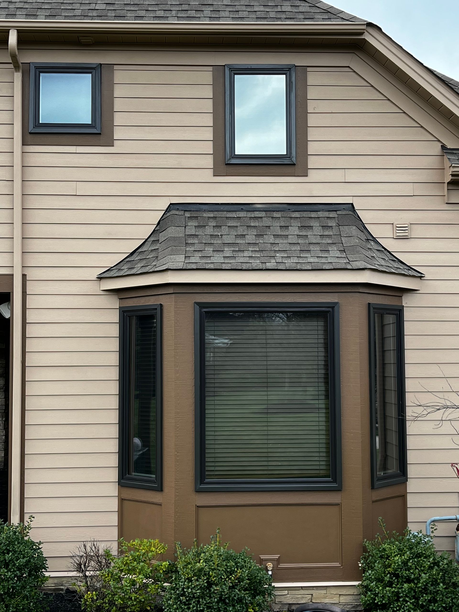 Beige house exterior with dark trim and windows, a bay window with a brown roof, and landscaping.