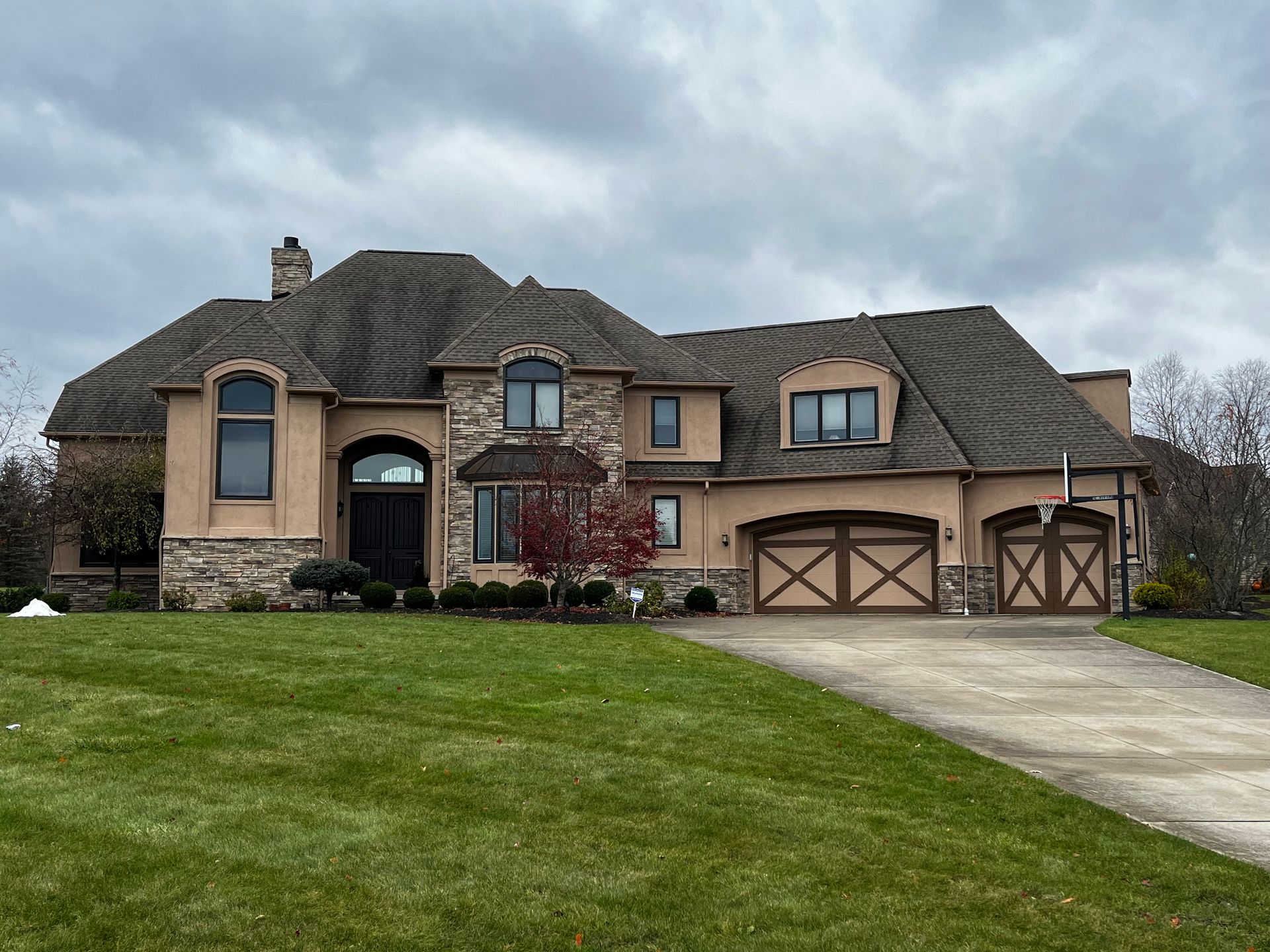Large tan and stone house with a multi-paneled garage door and a sloped, shingled roof under a cloudy sky.