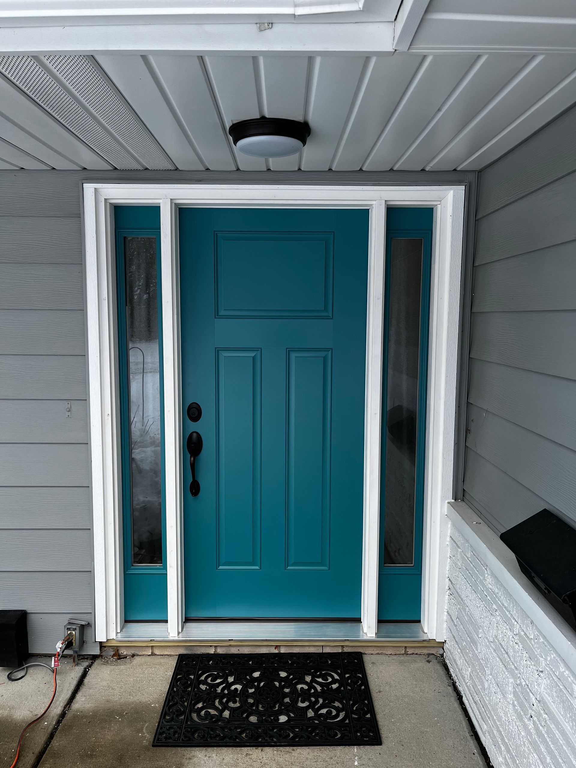 Teal front door with sidelights, white trim, under a gray porch ceiling, with a black mat.