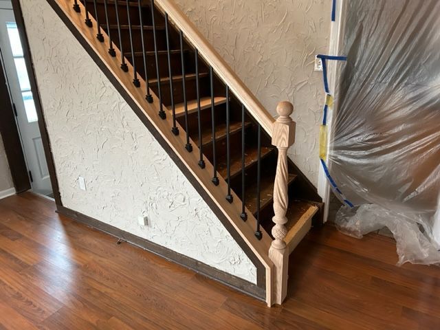 Staircase with light wood railing, dark spindles, and brown treads against textured white wall, over brown flooring.