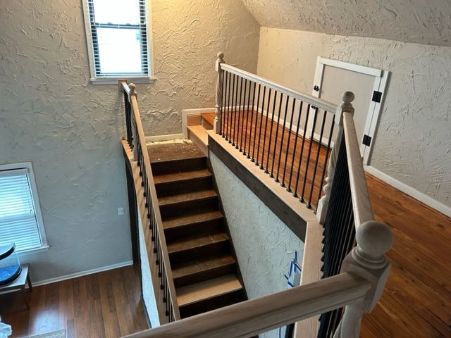 Staircase with wooden steps and railing leading upwards, against textured beige walls. Sunlight through a window.