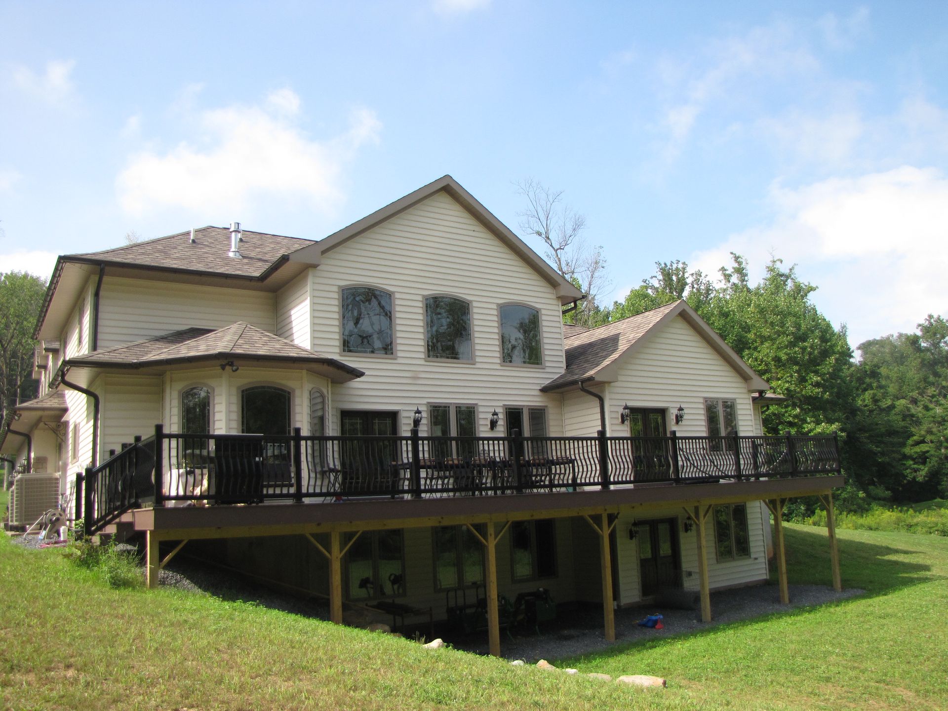 Two-story beige house with brown deck and black railing, set on a grassy hill under a blue sky.