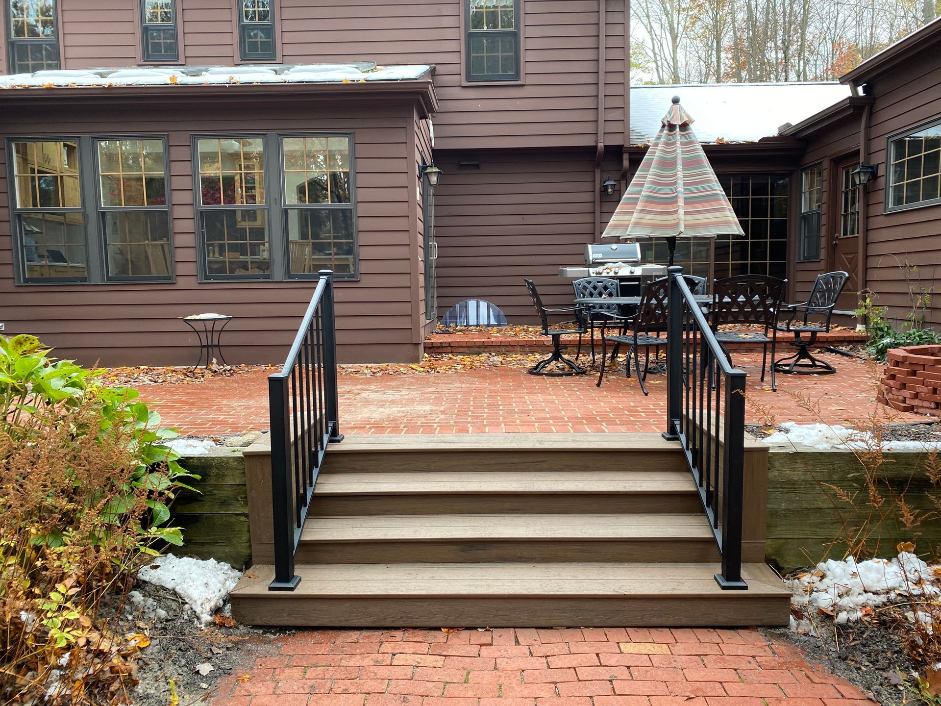 Brown house with brick patio, steps with black railings. Outdoor furniture and umbrella.