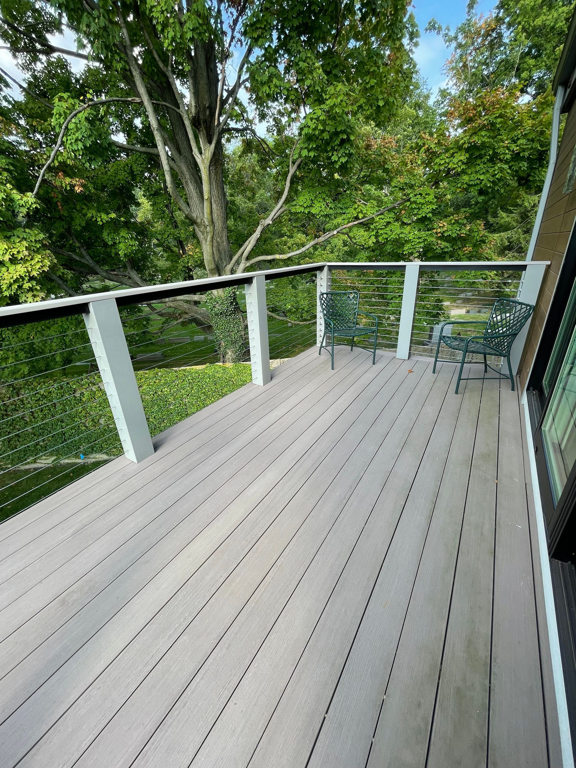Wooden deck with glass railing, two green chairs, and trees in the background.