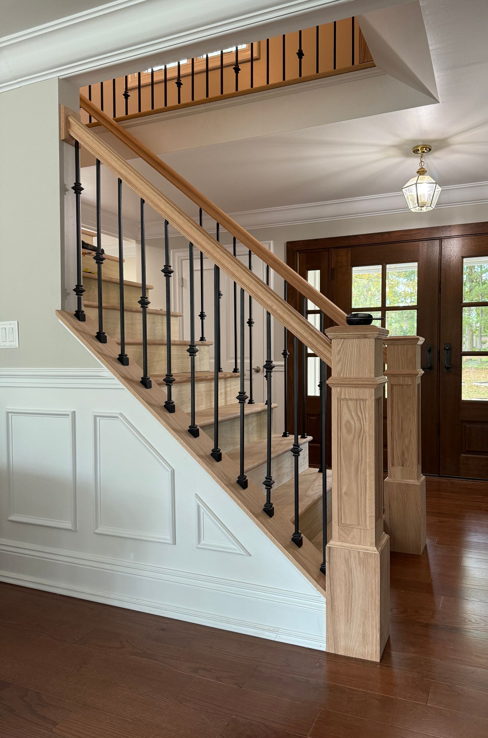 Staircase with light wood handrails and black balusters. A wooden door and light fixture are visible in the background.