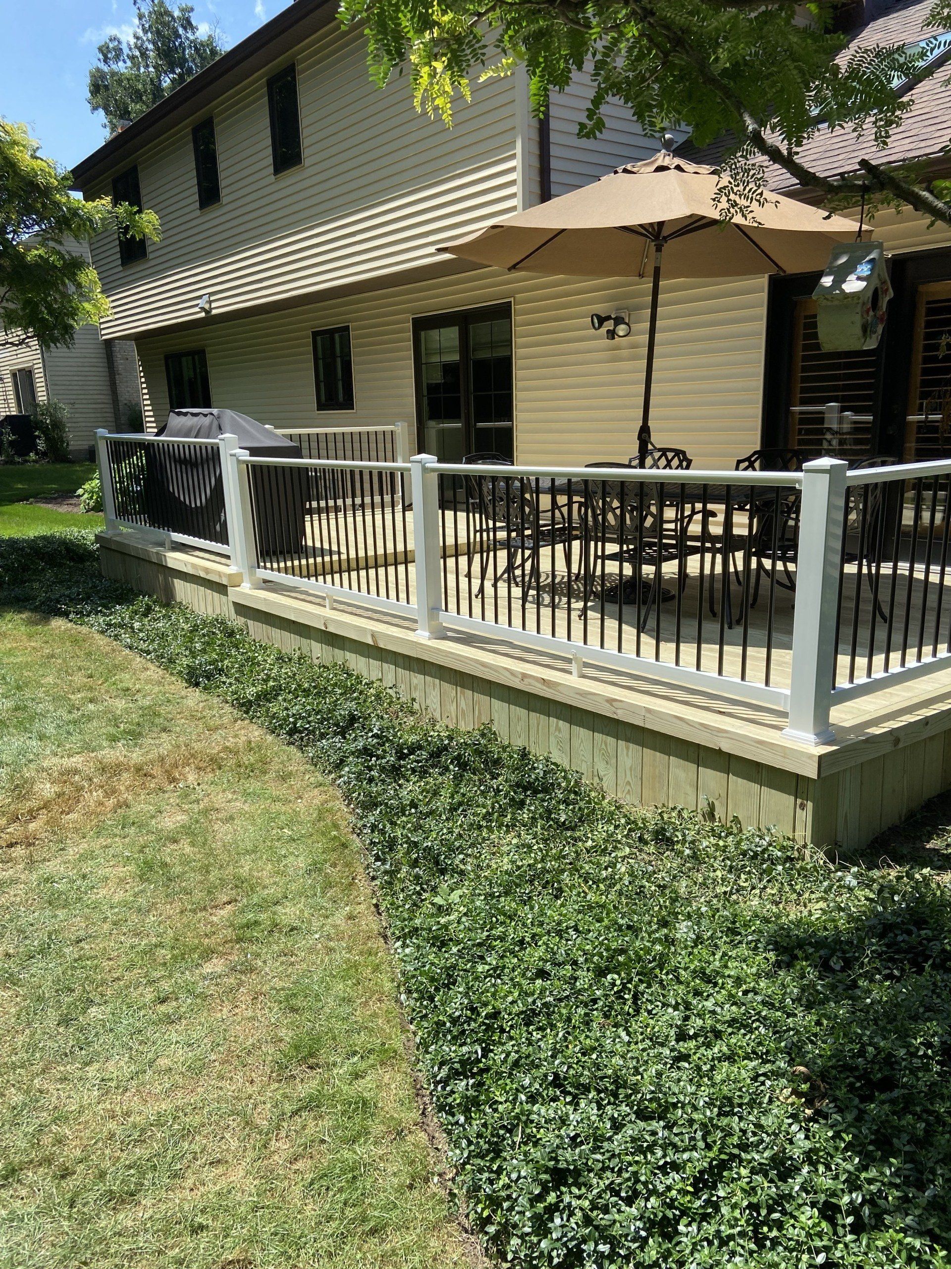 Backyard deck with railing, table, umbrella, and a house in the background. Green bushes and grass are in the foreground.