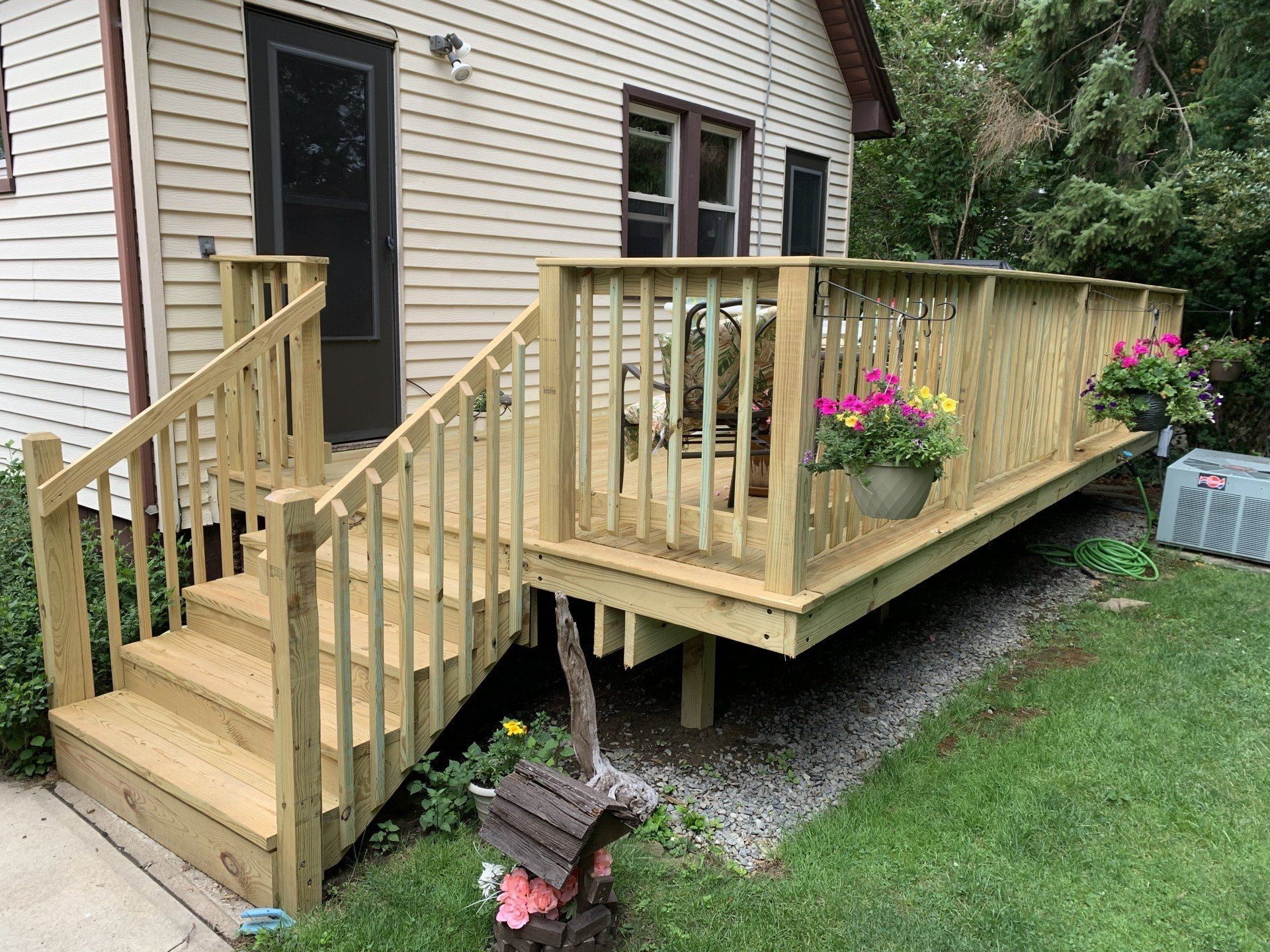 Wooden deck with stairs, built on the side of a beige house, with potted flowers and a seating area.