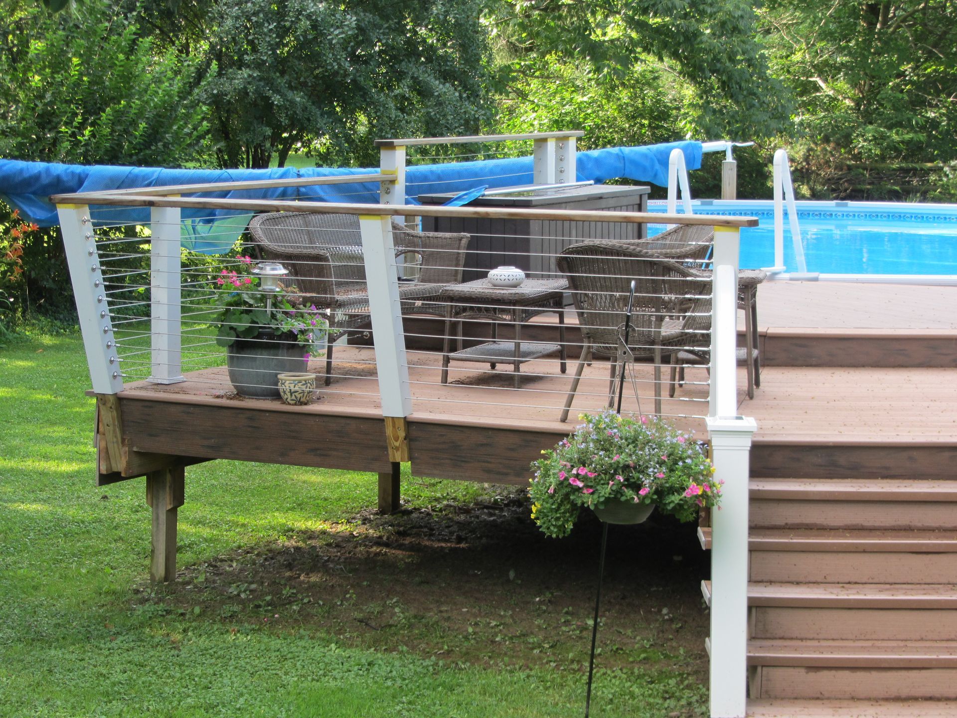 Wooden deck with patio furniture, overlooking a pool. White railing, green grass, and trees are also visible.