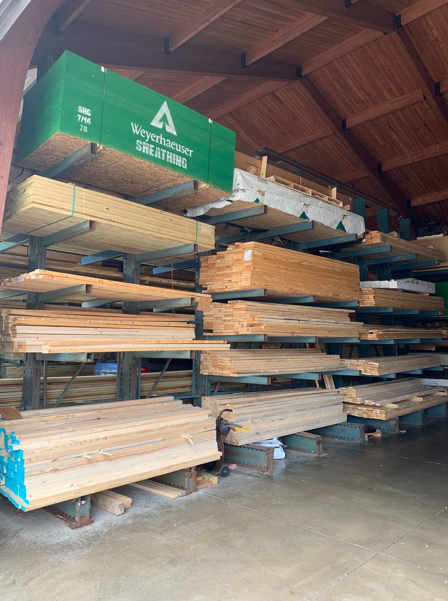 Lumber storage rack filled with various wood planks and boards inside a wooden building. Green box on top shelf.
