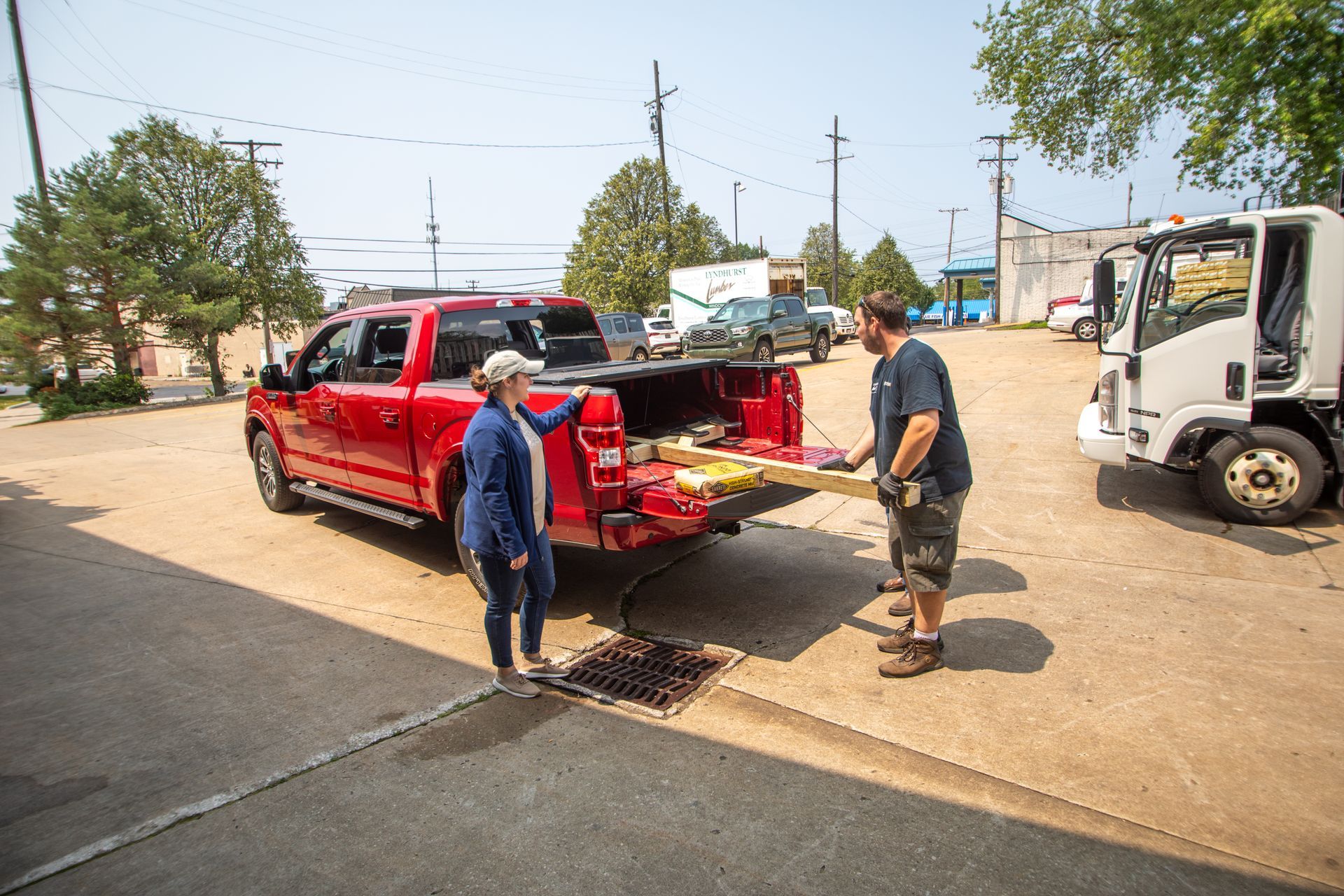A man and woman loading lumber into a red pickup truck. A white truck is parked nearby.