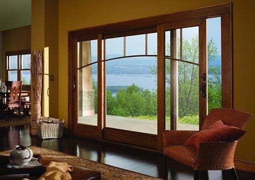 Wooden sliding glass doors open to a scenic view of a lake and trees. Interior features a wicker chair and table.