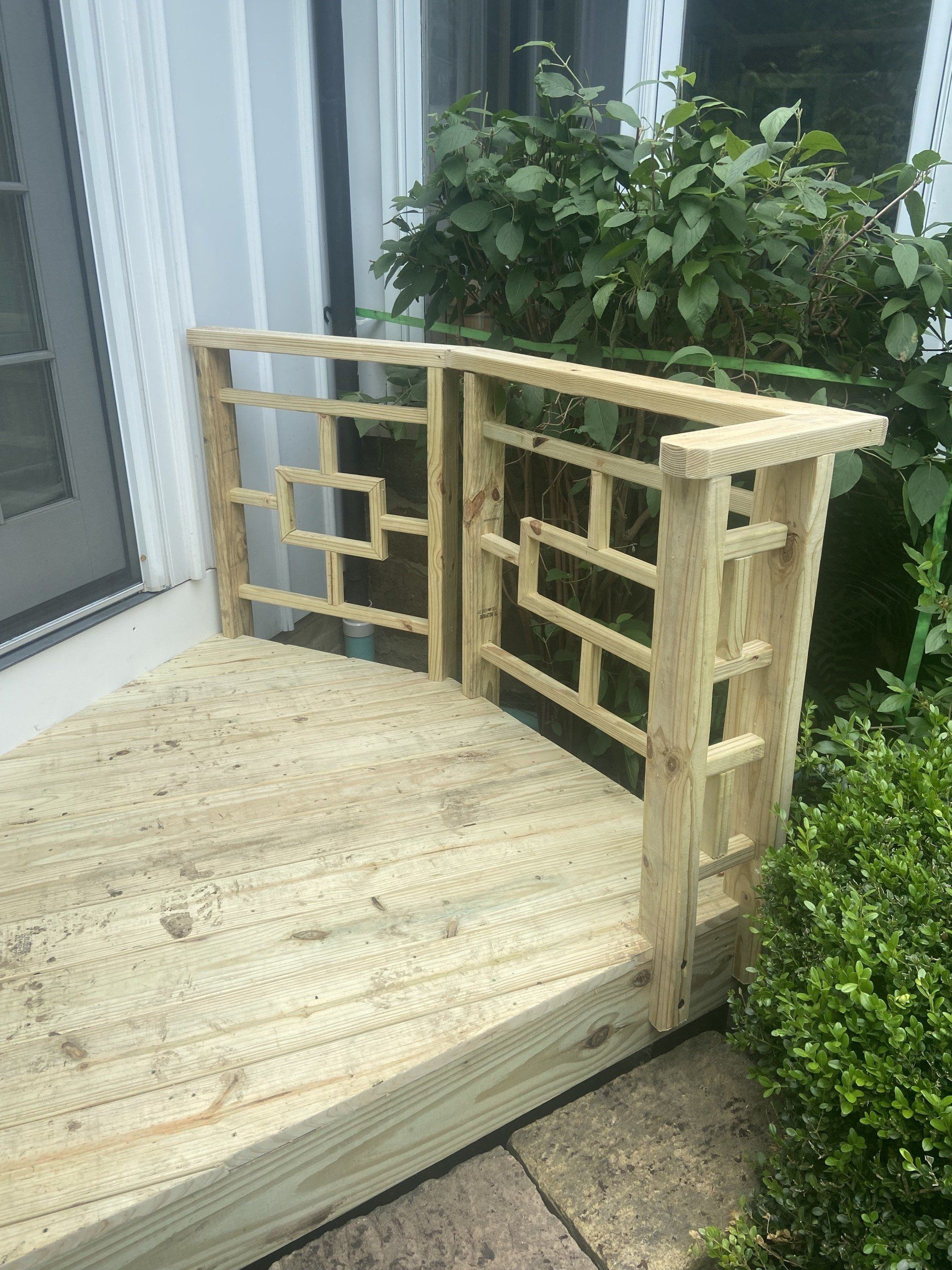Wooden deck with decorative railing, beside a door, next to shrubs.