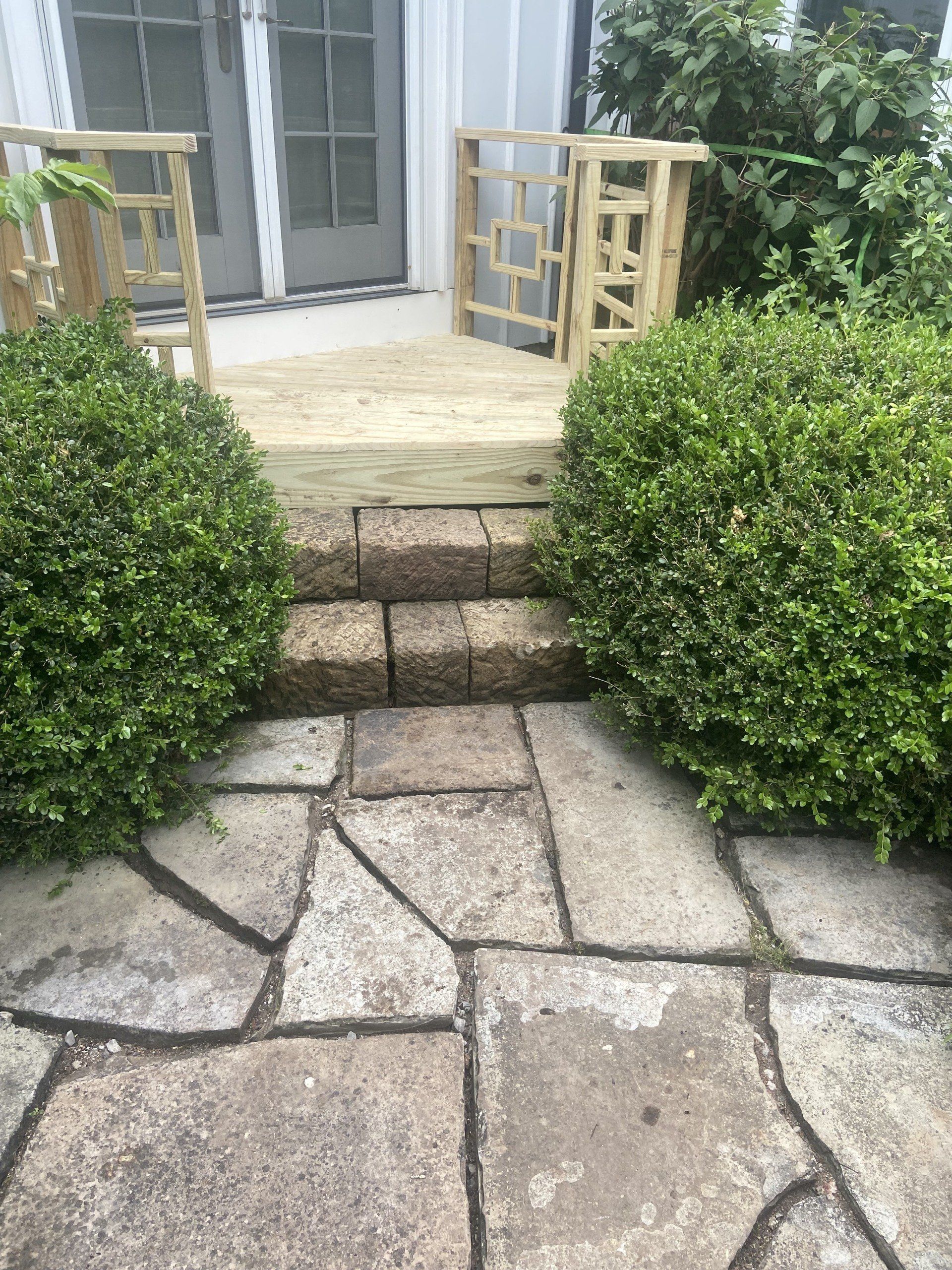 Stone steps leading to a wooden deck with bushes on either side. A door is visible in the background.