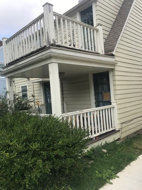 Beige two-story house with a porch and balcony. Green bushes and grass in the front.