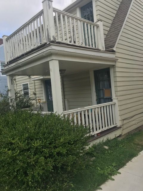 Tan two-story house with balcony and porch, white railings, and green bushes in front.