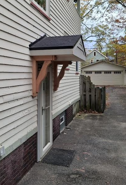 A door with a black-roofed wooden awning, mounted on a white house's siding, with a dark mat.