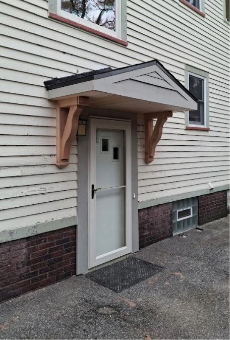 Tan-and-black awning above a white door set in a light-yellow building with a brick base.