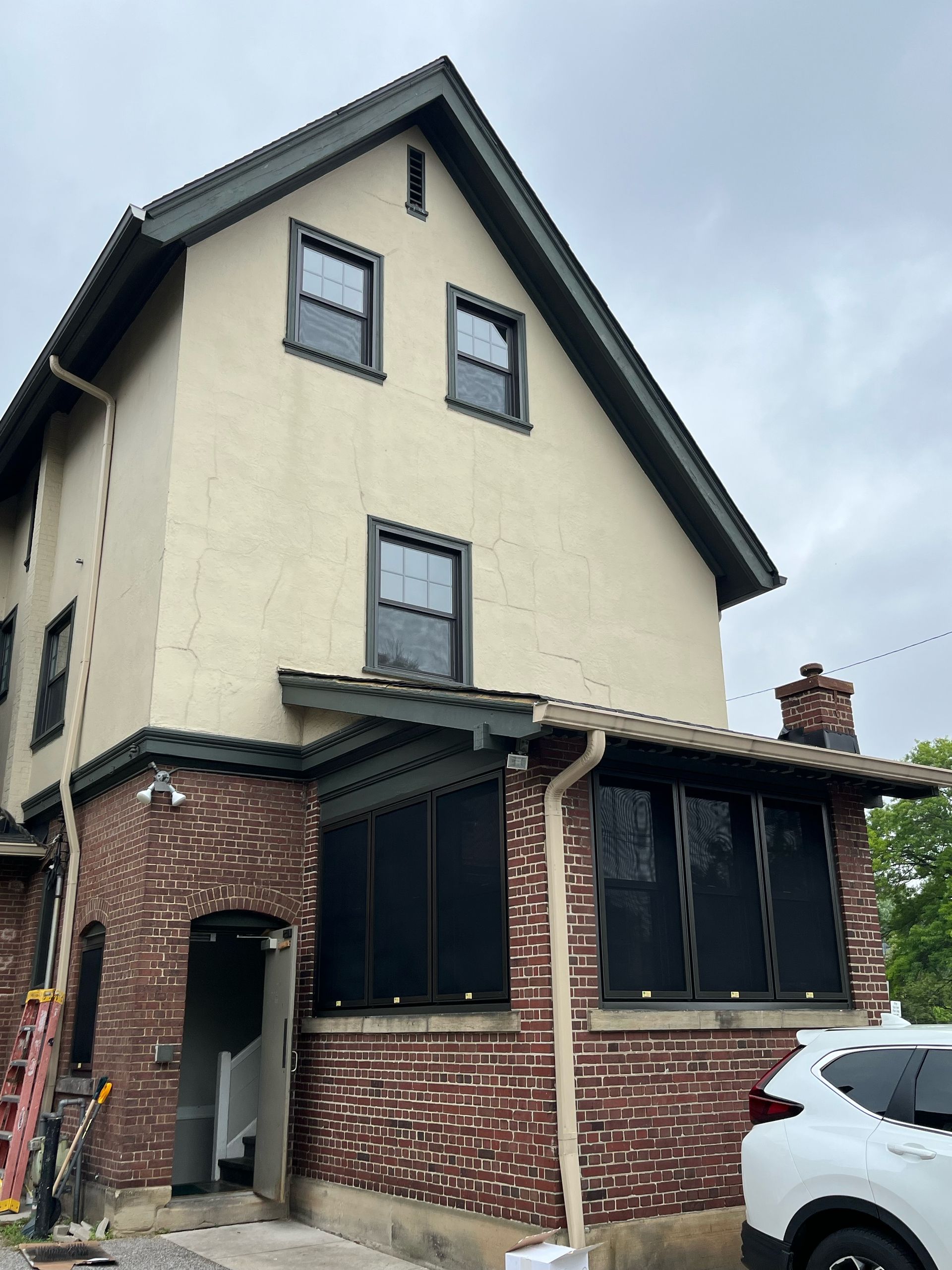 Three-story house with tan stucco, brick, and dark green trim, under overcast sky. A white SUV is parked nearby.